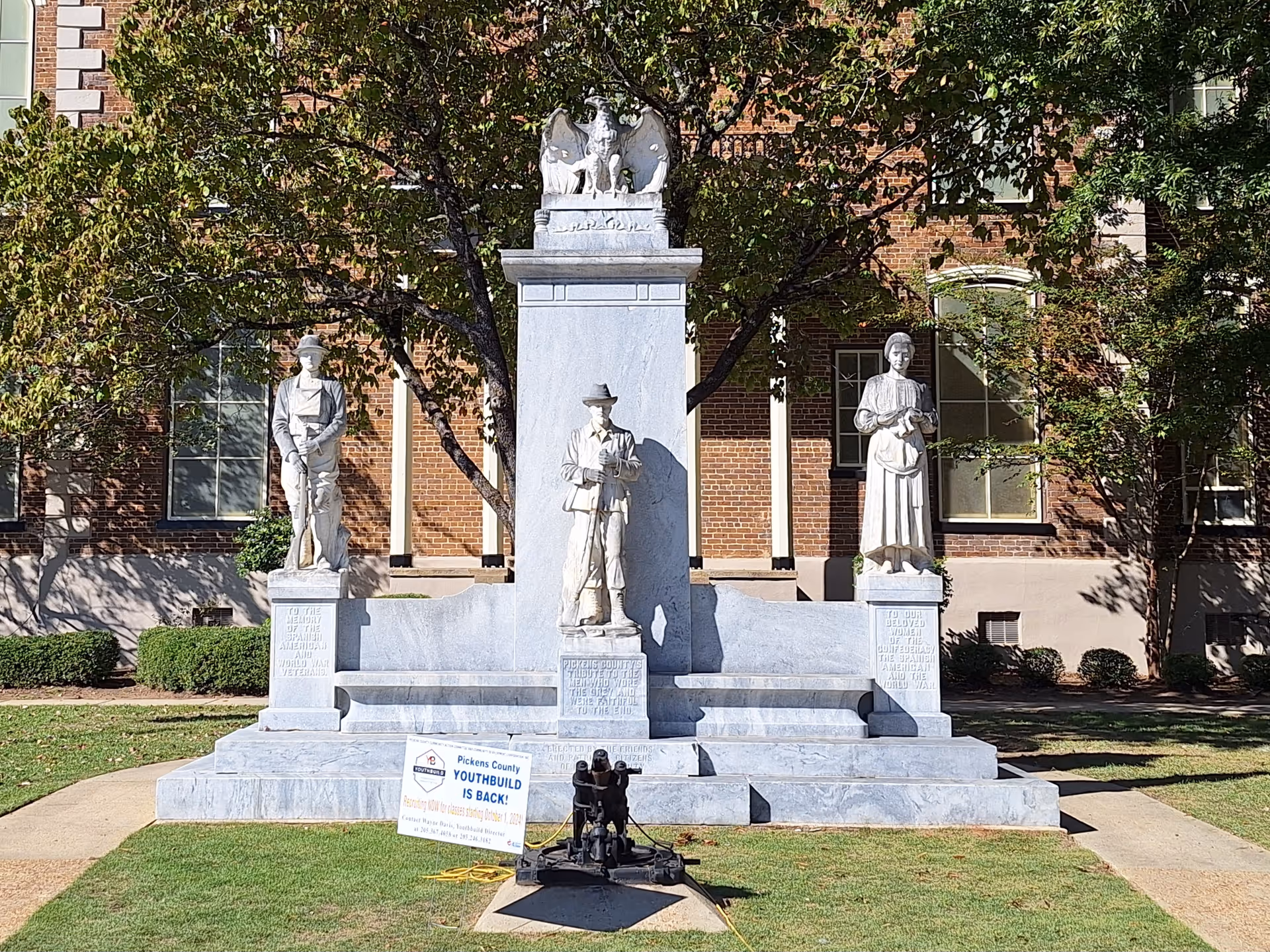 Stone monument with statues on a grassy lawn in front of a brick building.