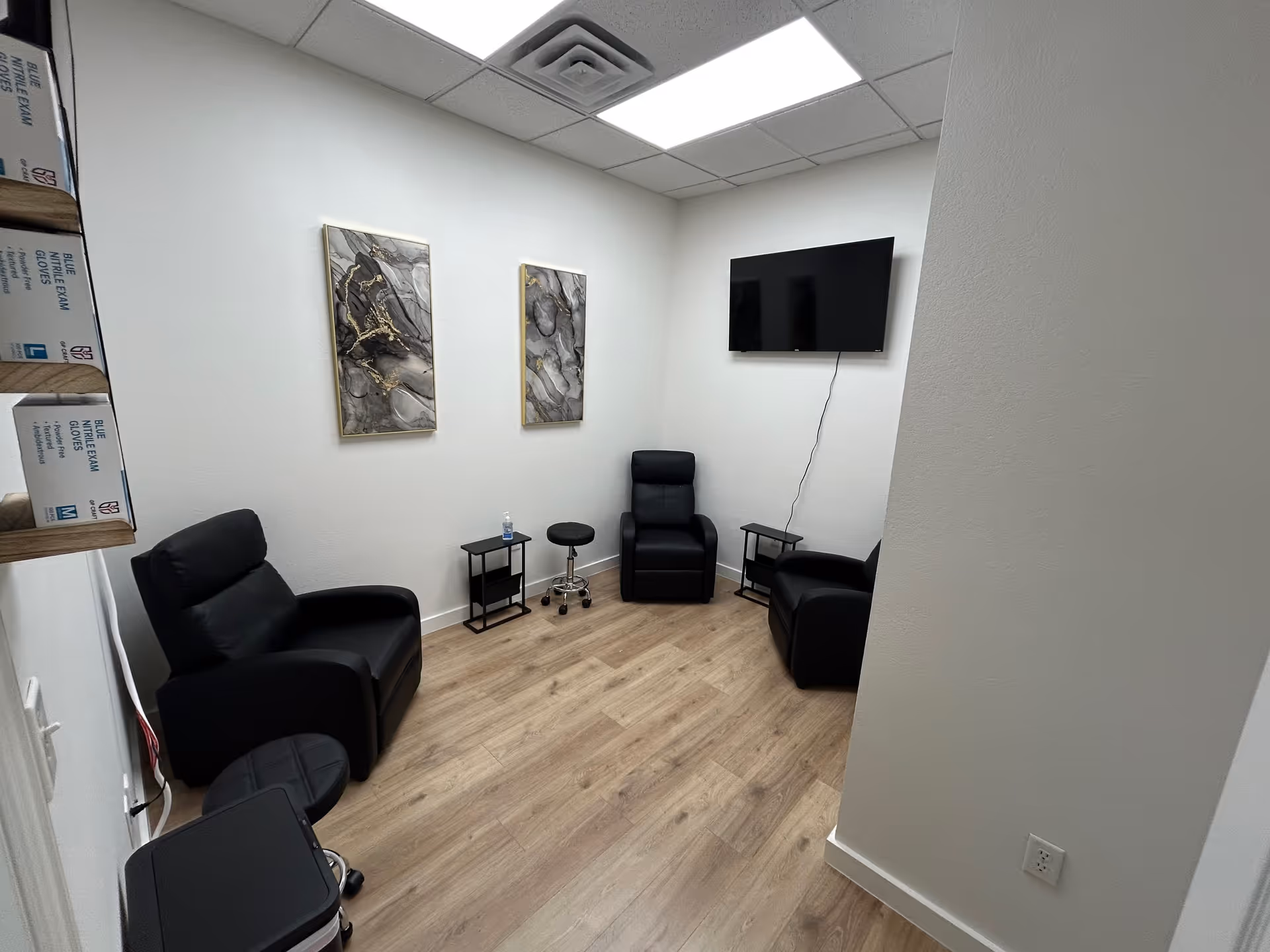 Small waiting area with black leather chairs, glass tables, wall art, and a mounted TV.