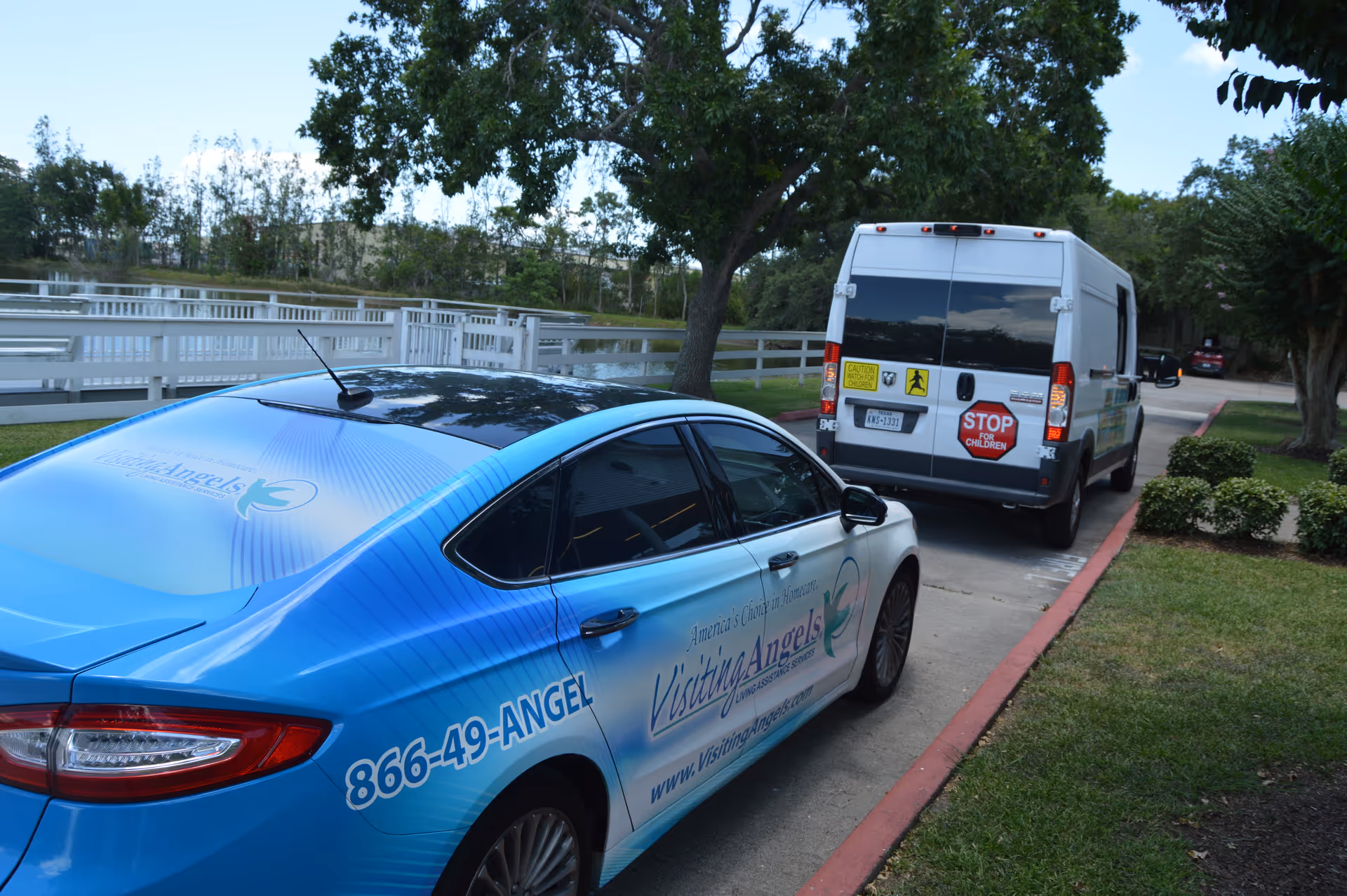 Blue Visiting Angels car and white van parked along a tree-lined outdoor driveway.