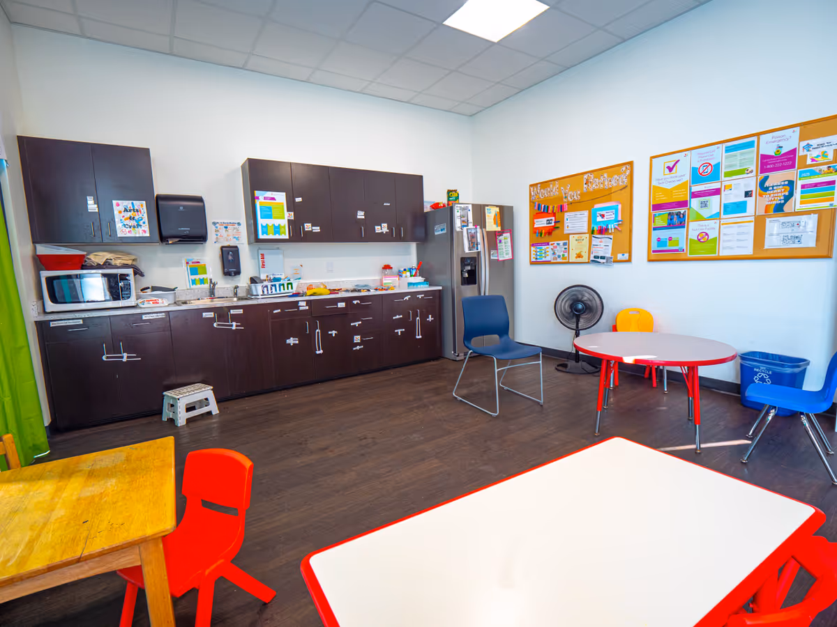 Bright communal kitchen area with dark cabinets, fridge, microwave, and child-sized tables and chairs.