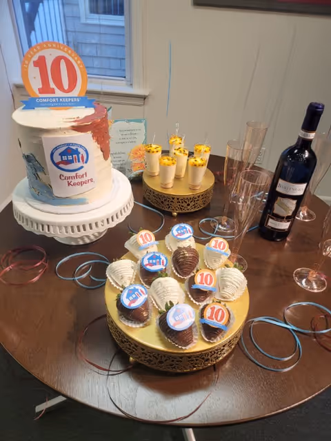 Celebration setup on a dining table with a Comfort Keepers cake, cupcakes, wine bottle and ribbons.