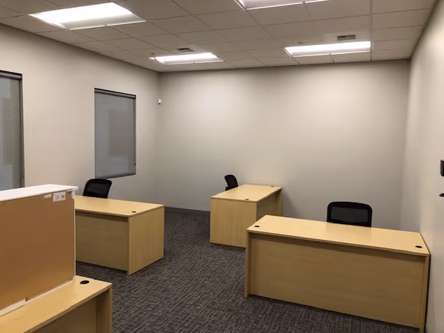 Office space with several wooden desks, black chairs, gray carpet, and bare walls.