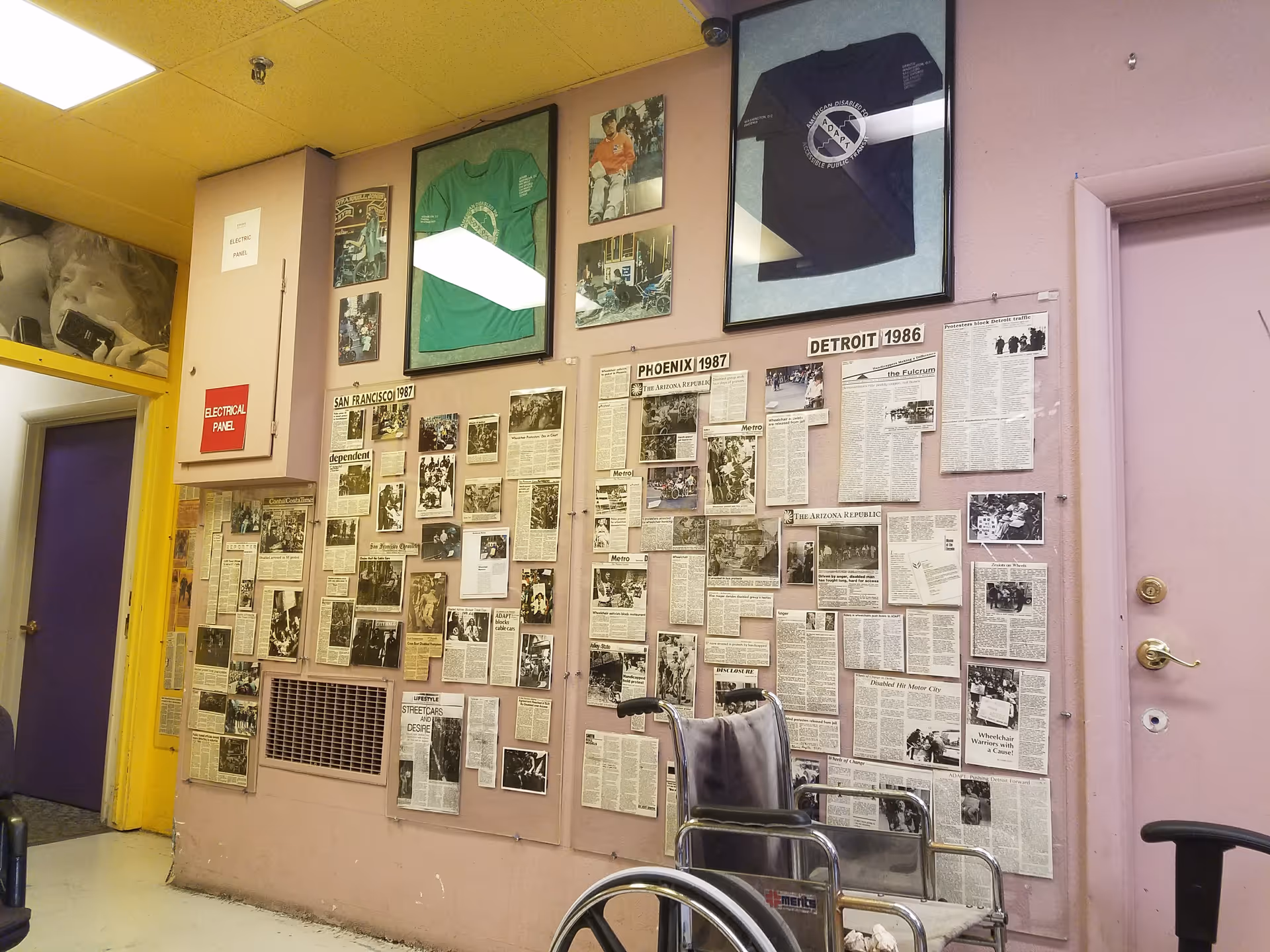 Pink-walled interior hallway with newspaper clippings display and a wheelchair in foreground.