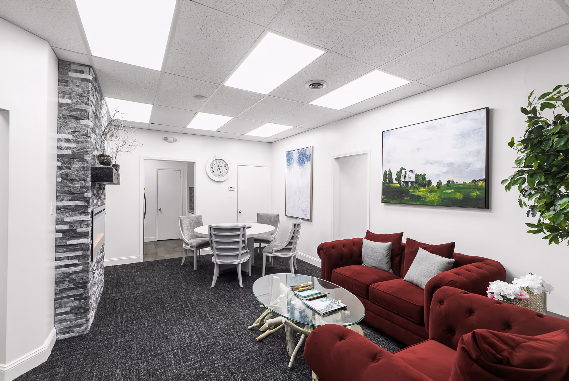 Spacious senior living lounge with red sofas, glass coffee table, paintings, and stone fireplace.
