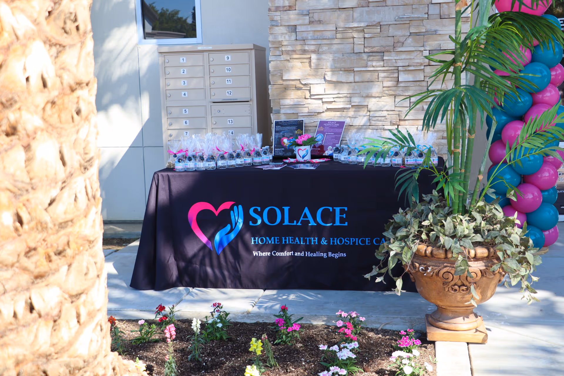 Outdoor table with Solace Home Health & Hospice Care banner and balloons beside potted plants.