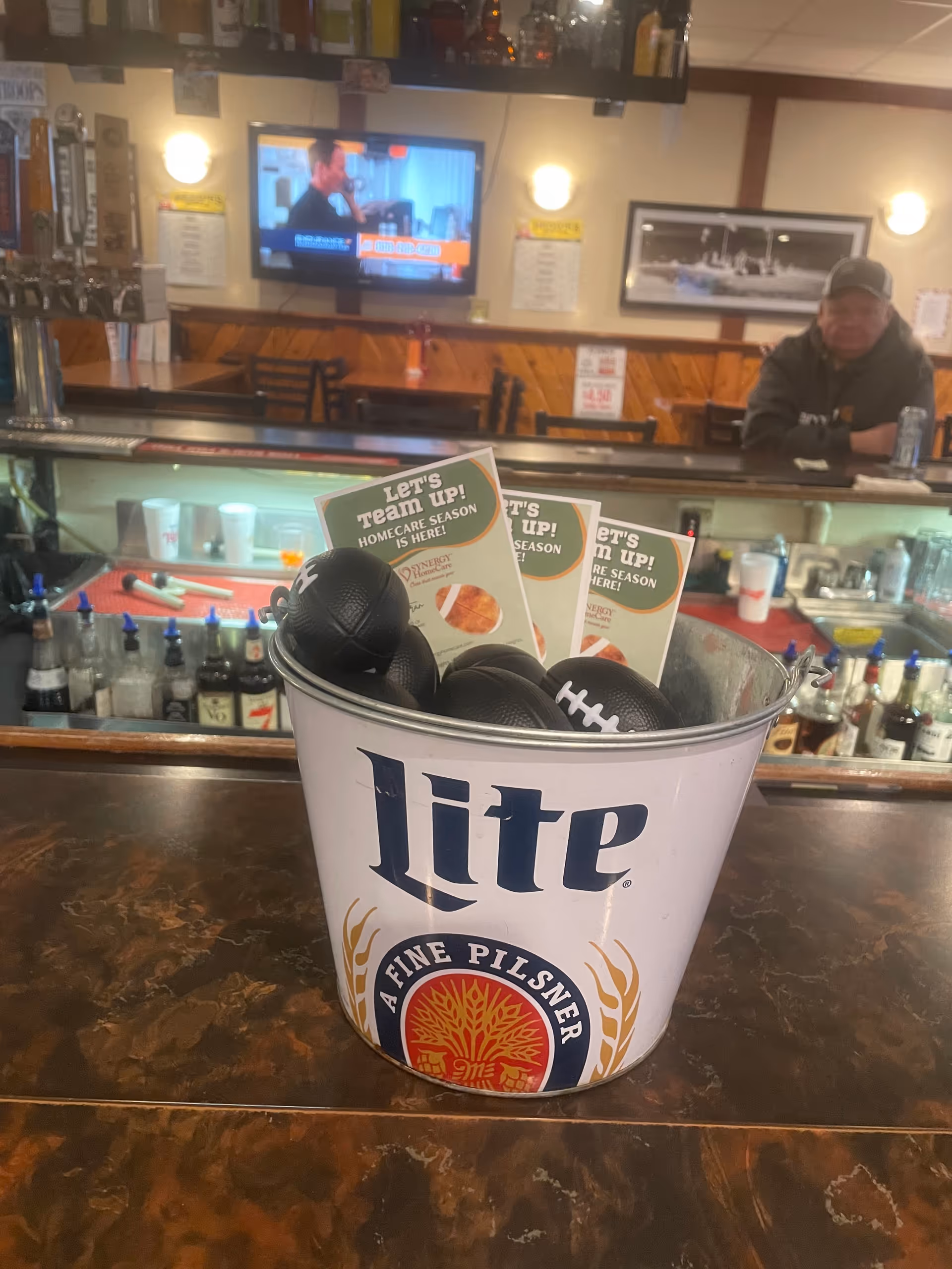 A bar counter with a Lite beer bucket and promotional flyers, man seated in background.