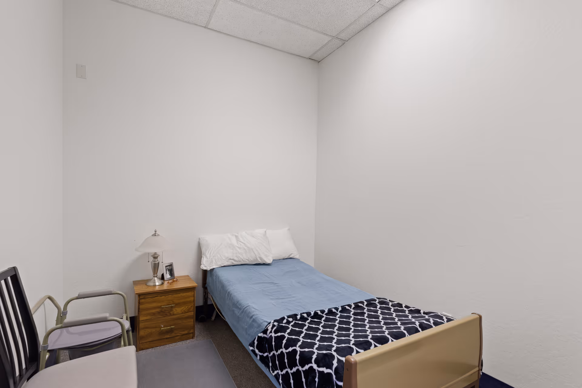 Small bedroom in a senior living facility with a bed, nightstand, lamp, and chair.