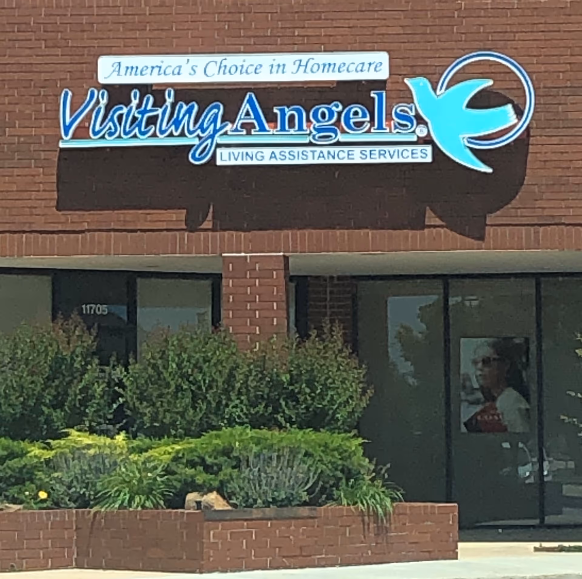 Red-brick building facade with a large Visiting Angels sign and shrubs.