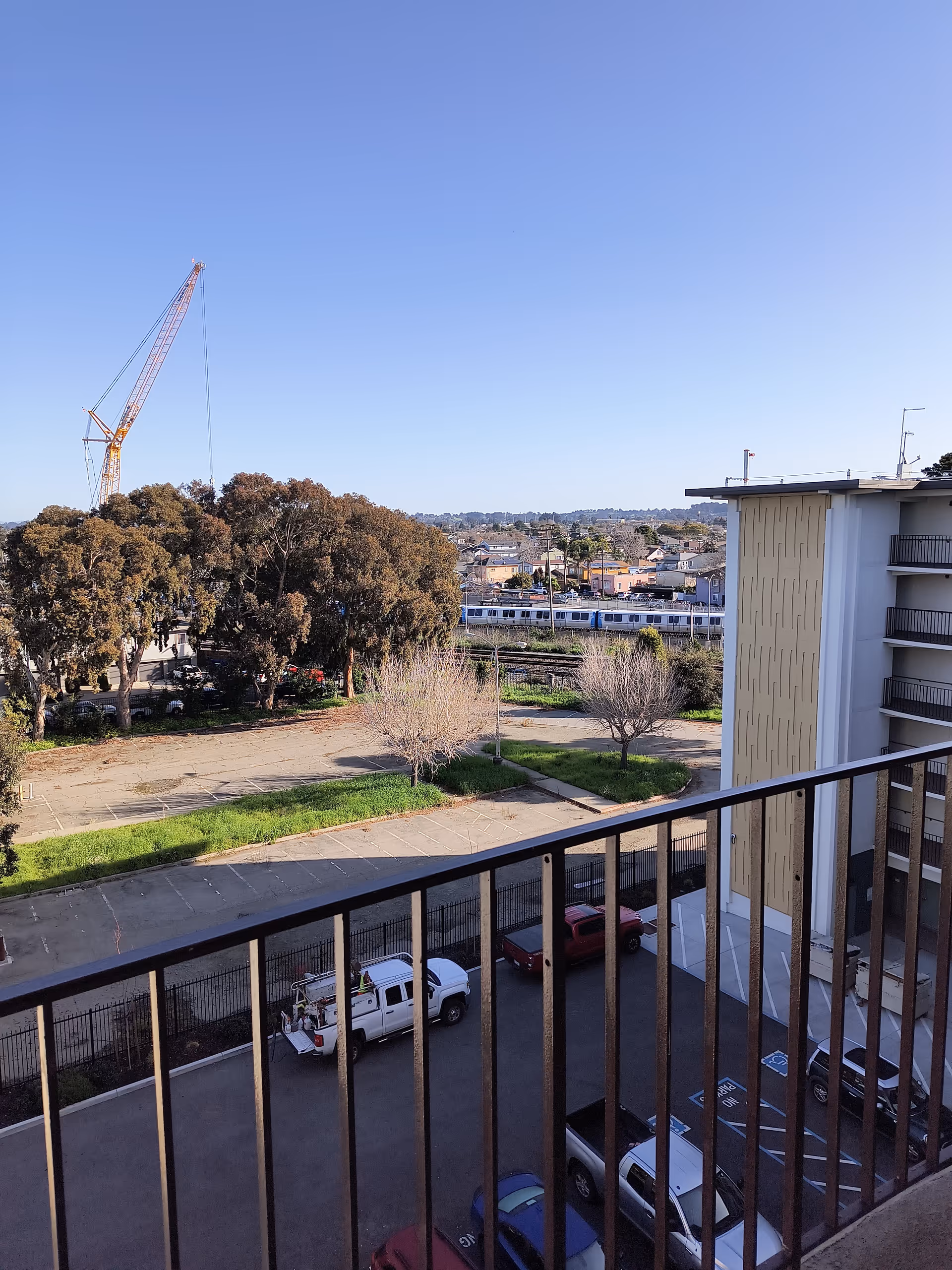 Balcony railing overlooks a parking lot, trees, and distant construction crane against a clear blue sky.