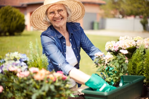 Smiling elderly woman wearing a sunhat tends flowers outdoors at FirstLight Home Care of Warrenton.