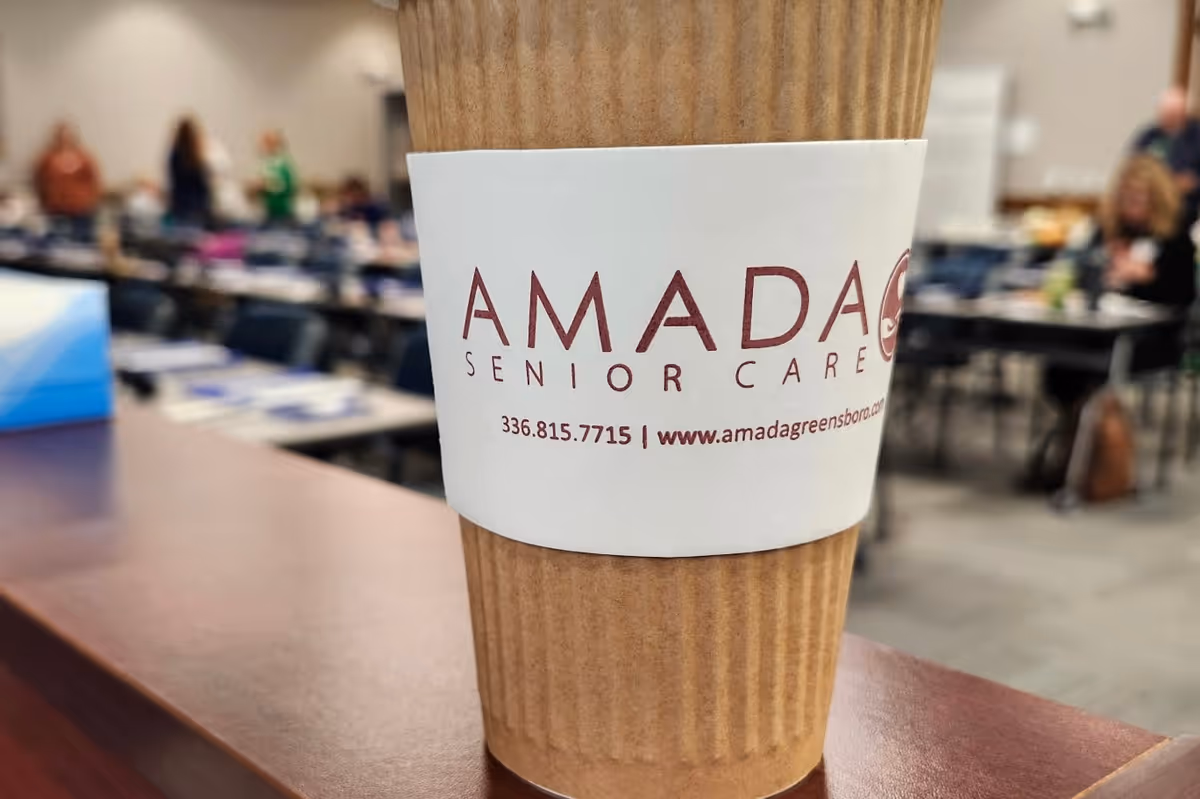 Close-up of a coffee cup sleeve reading Amada Senior Care with a busy meeting room behind.