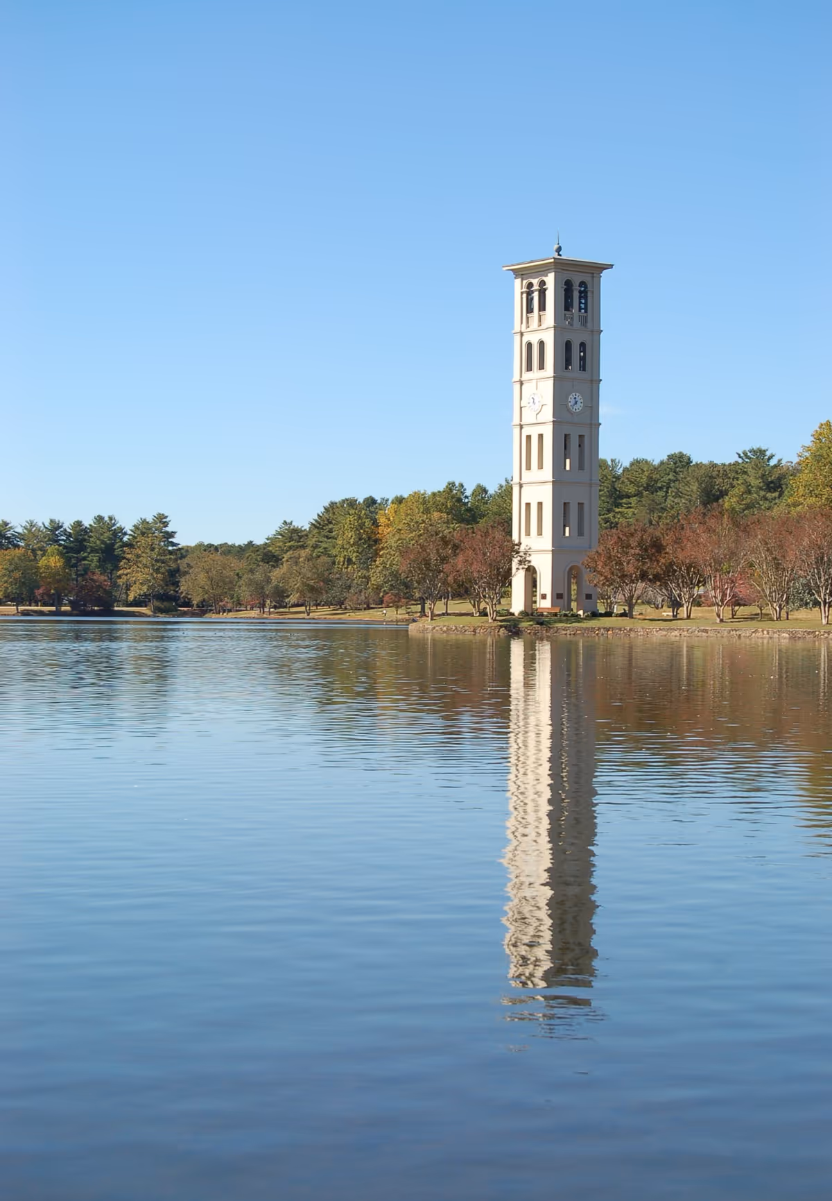 Tall clock tower reflected in calm lake with trees in a park setting.