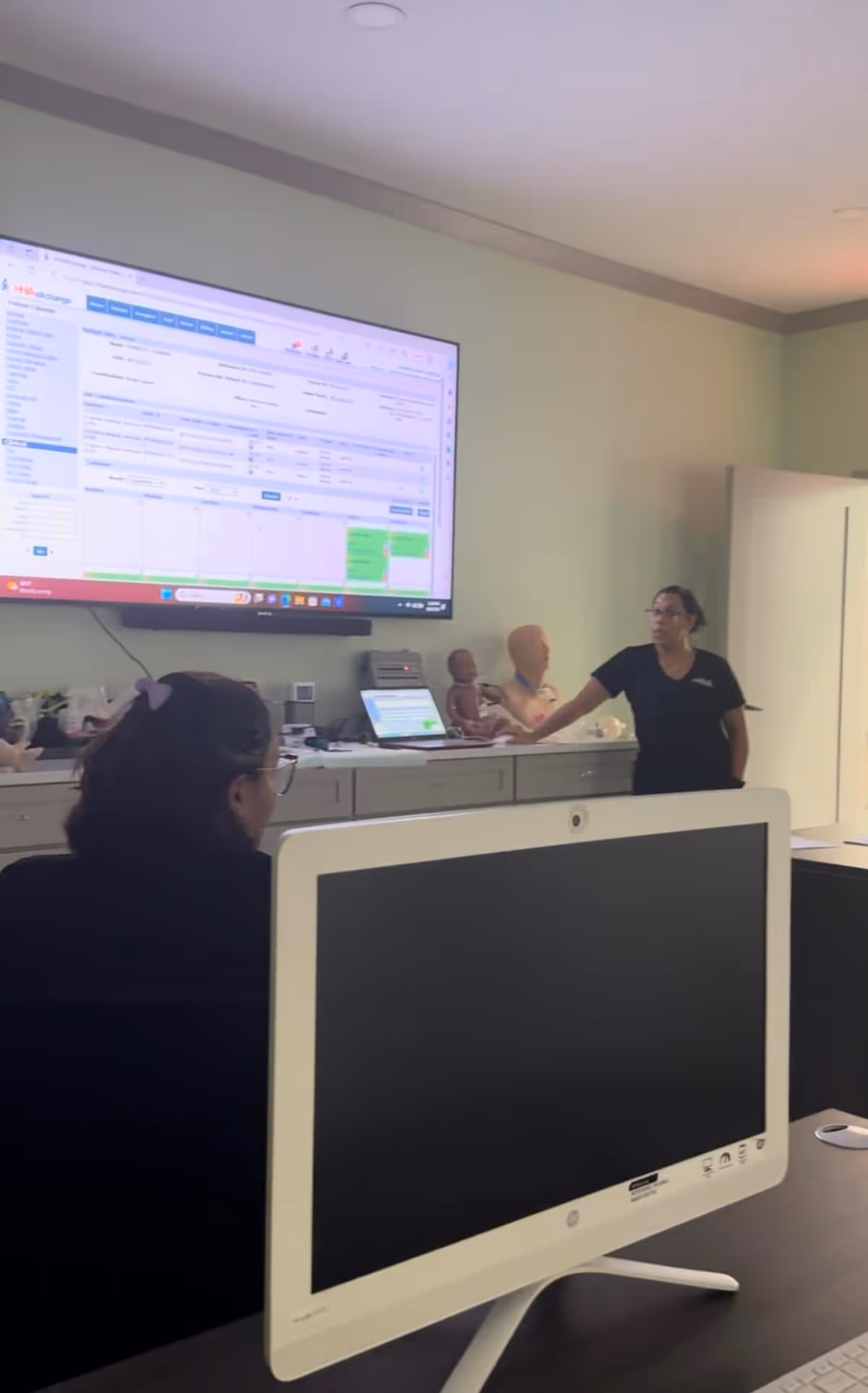 A presenter stands near a wall-mounted monitor in a training room with mannequins.
