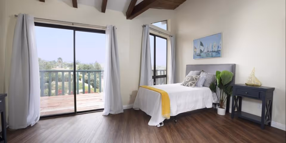 A bright bedroom with a bed, sliding balcony doors, and wood flooring.