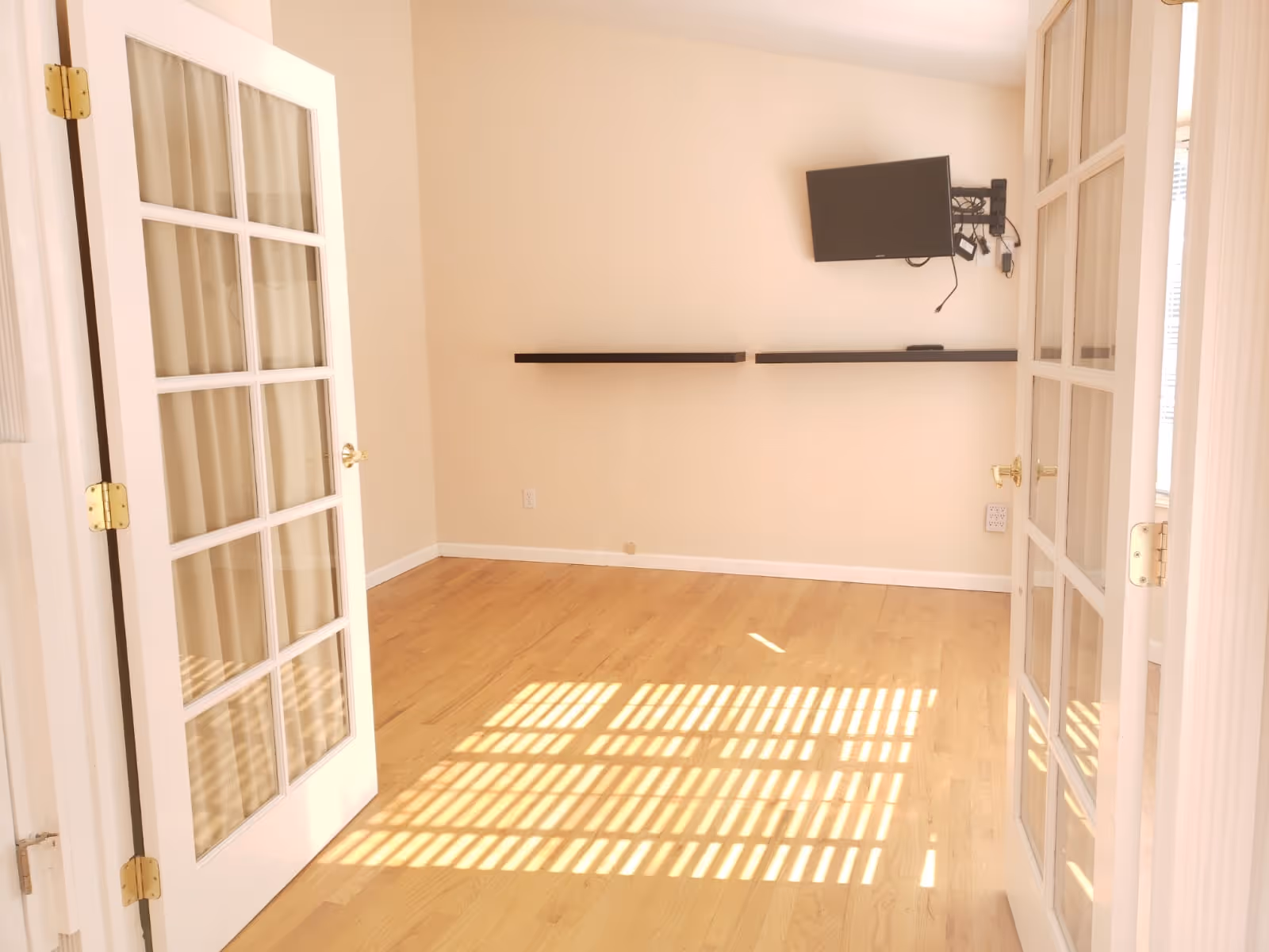 Empty interior room with French doors, wooden floor, wall-mounted TV, and shelves.