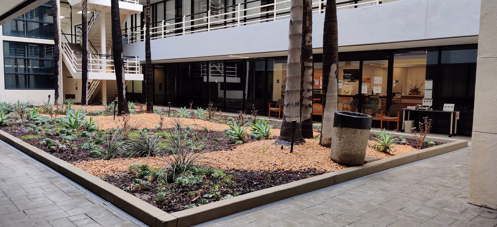 Outdoor courtyard with palm trees, landscaped beds, and a building facade with lobby seating.