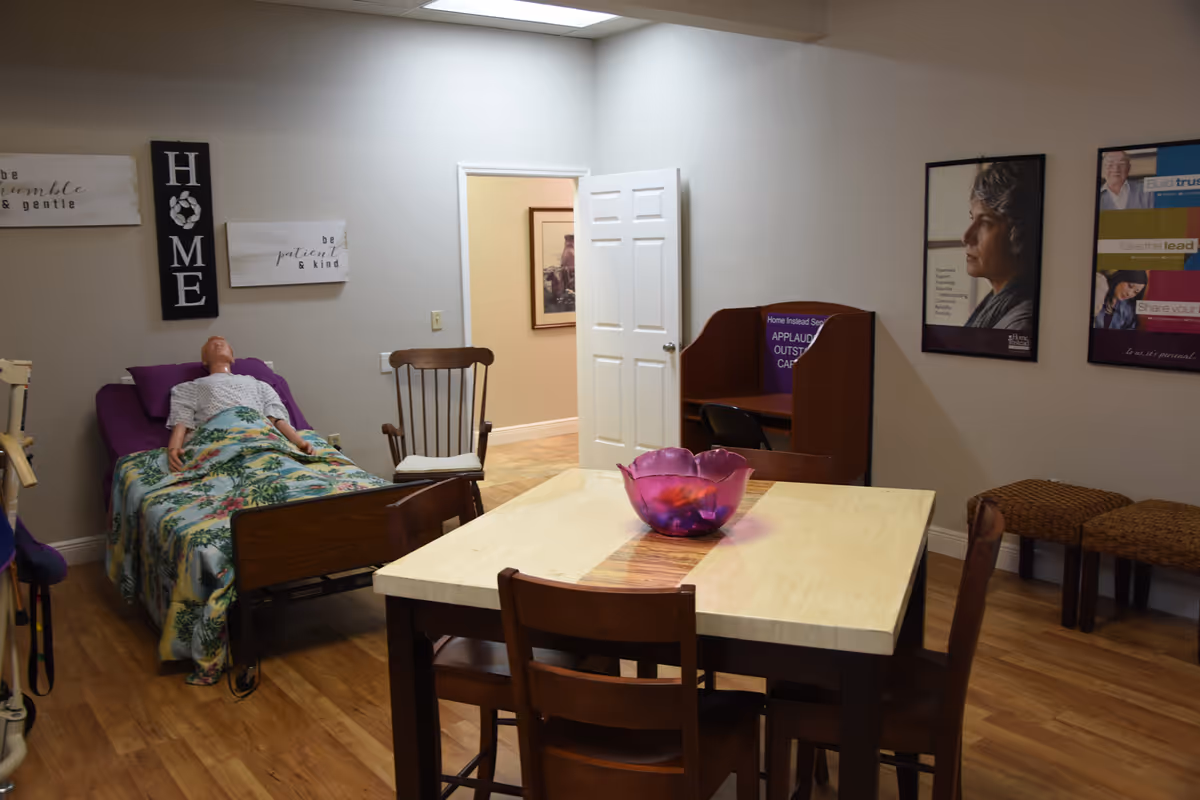 An elderly resident rests in a bed beside a dining table in a calm facility room.