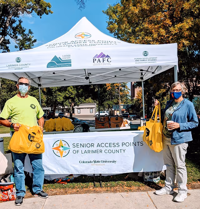 Outdoor outreach booth with masked volunteers and yellow tote bags beside a branded tent.