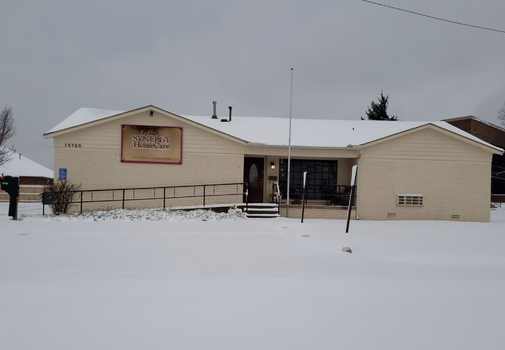 Single-story beige building with a SYNERGY HomeCare sign, snow-covered ground, and steps.