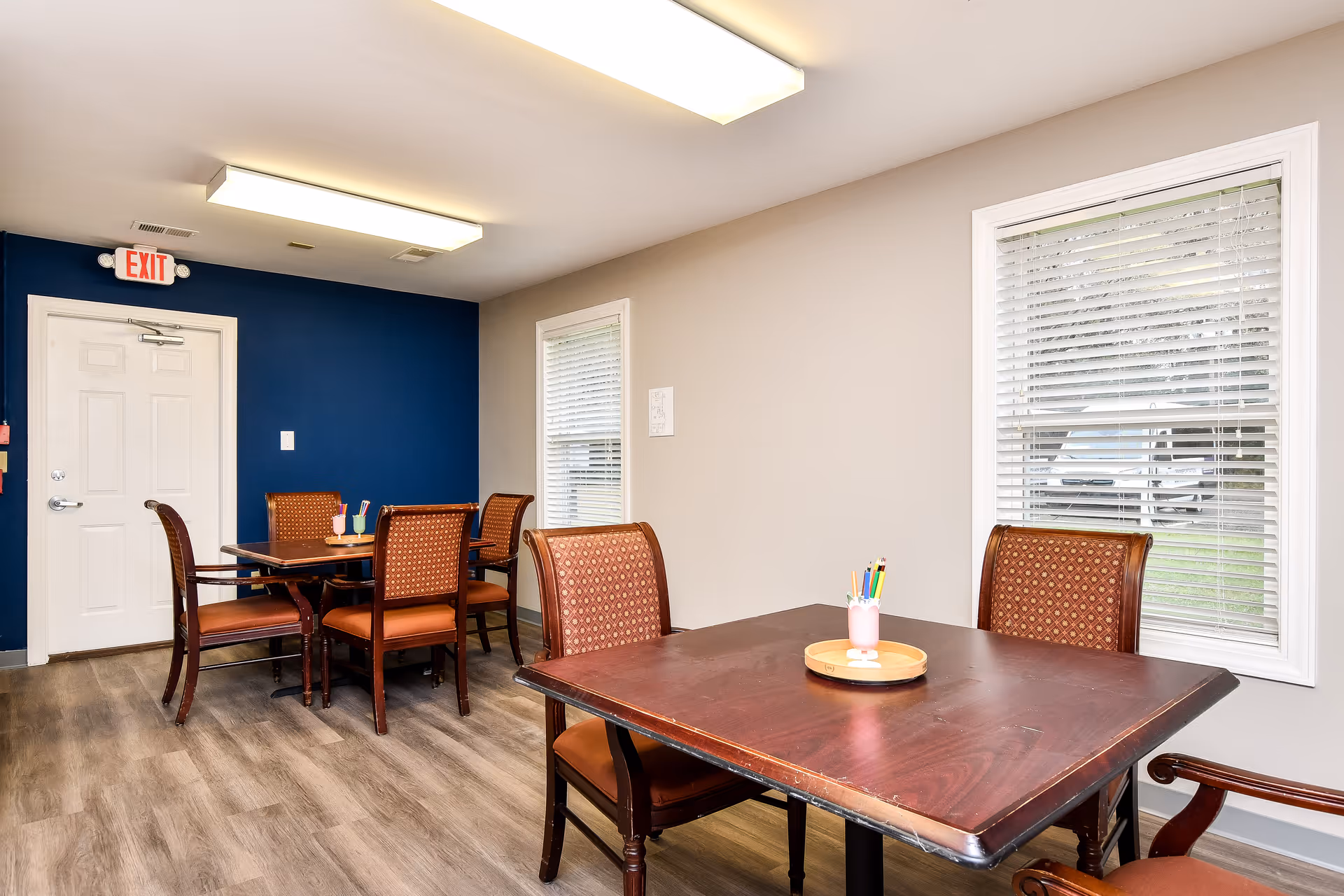 Communal dining room with wooden tables, chairs, windows, and an EXIT sign by a blue wall.