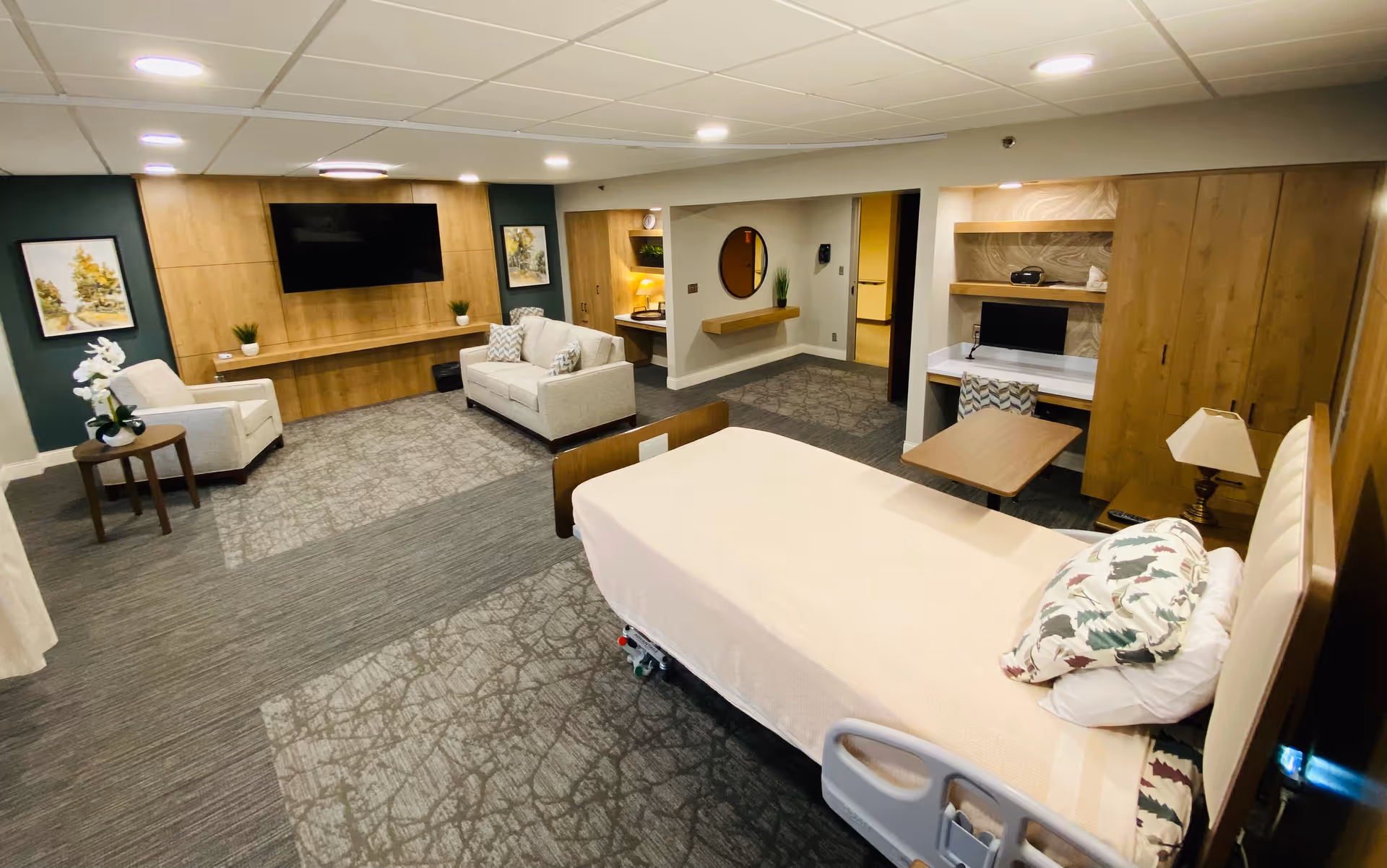 Interior hospice suite with a hospital bed, seating area, TV, and wooden cabinetry.