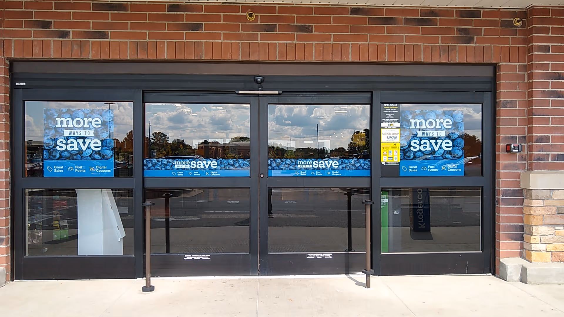 Brick storefront with glass doors and blue 'more ways to save' posters.