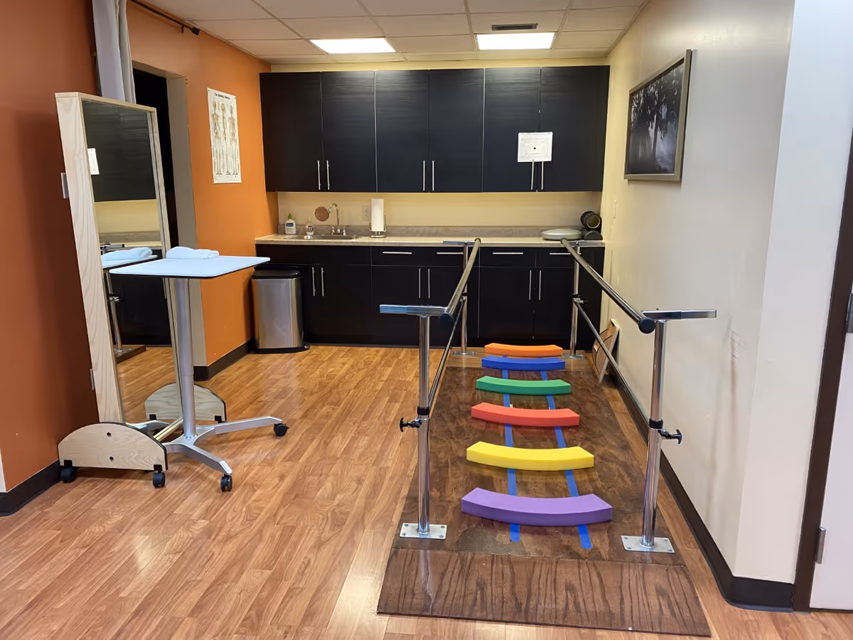 Therapy room with colorful stepping stones, parallel bars, and dark cabinetry kitchenette area.