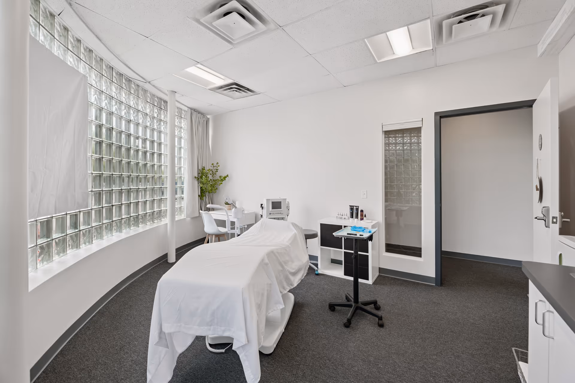 A clinical treatment room with a covered bed, medical equipment, and curved glass block window.