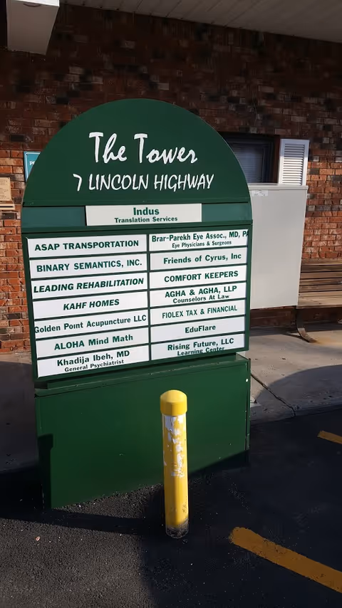 A green directory sign on a brick building exterior with a yellow bollard in front.