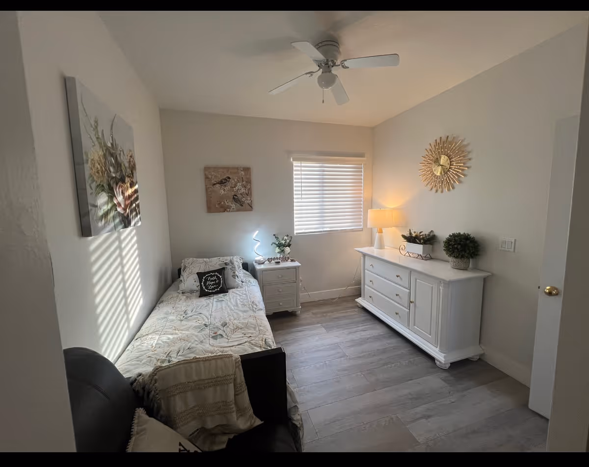 A small, neatly furnished bedroom with a bed, dresser, nightstand, and ceiling fan.