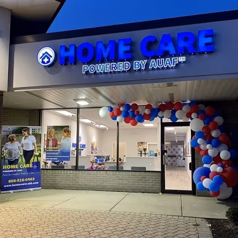 Storefront of Home Care Powered by AUAF with blue illuminated sign and balloon arch at entrance.