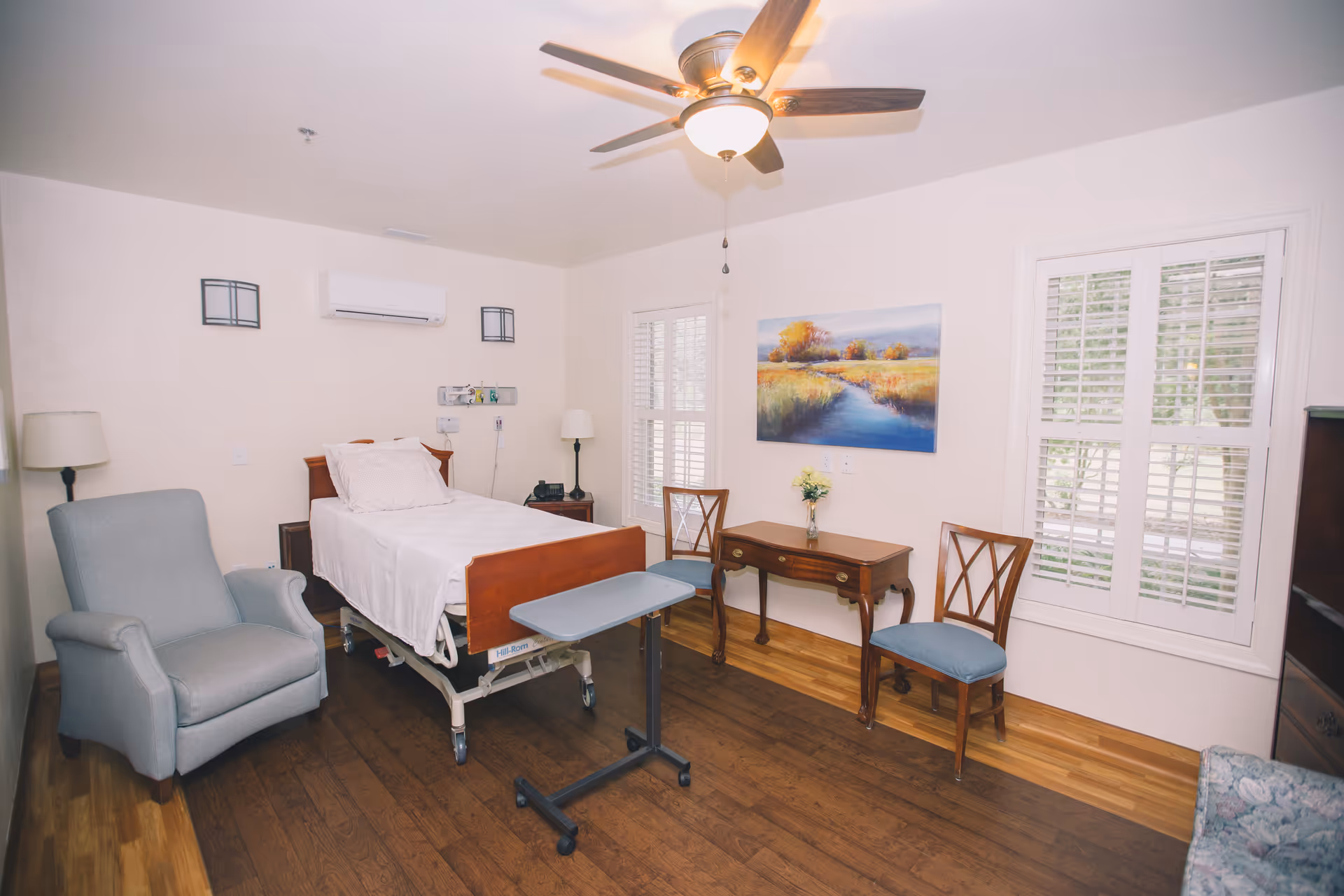Hospice bedroom with adjustable bed, blue chair, desk, and large windows.
