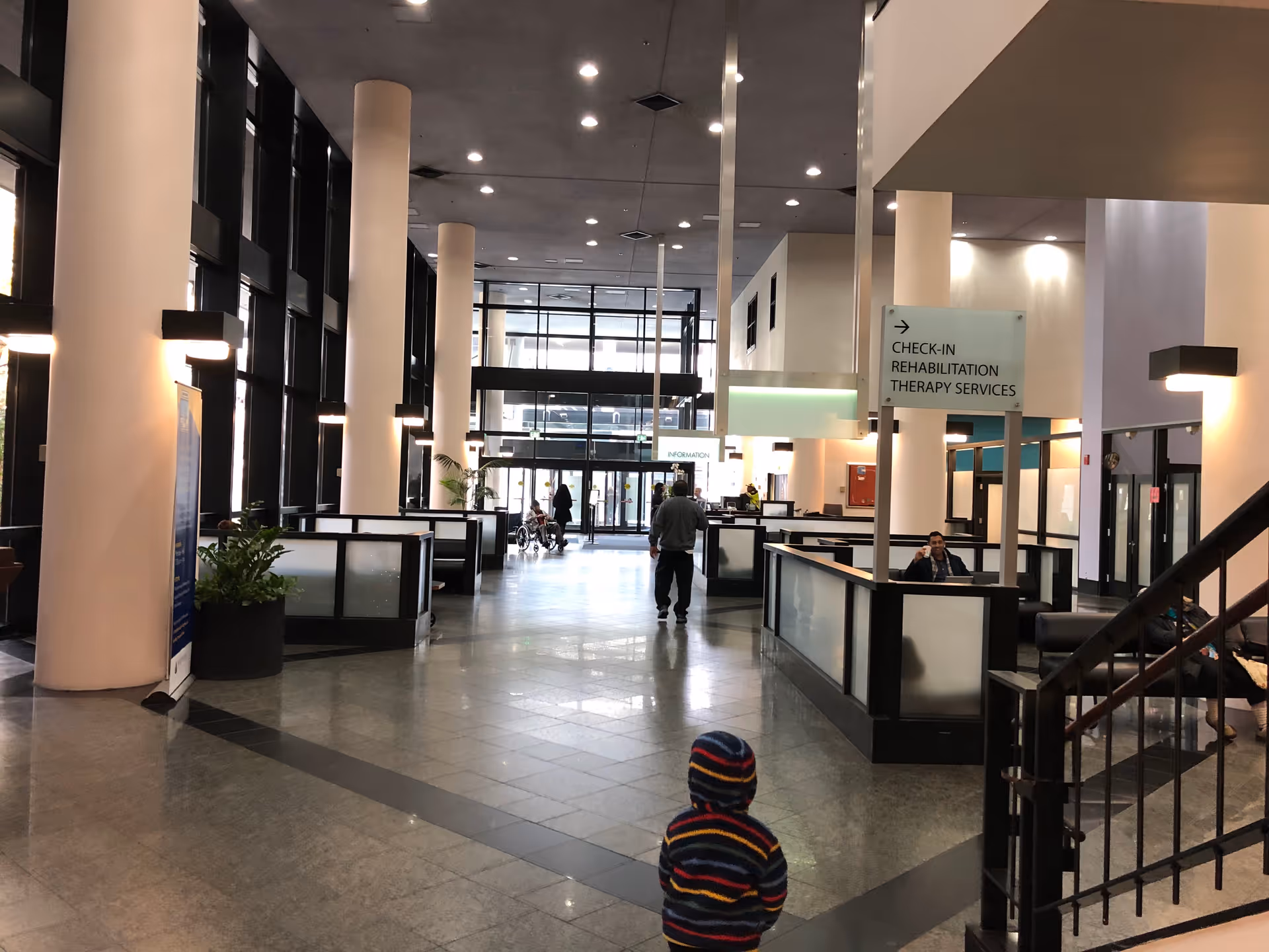 Hospital lobby with check-in desks, glass partitions, and a child in striped hoodie facing the area.