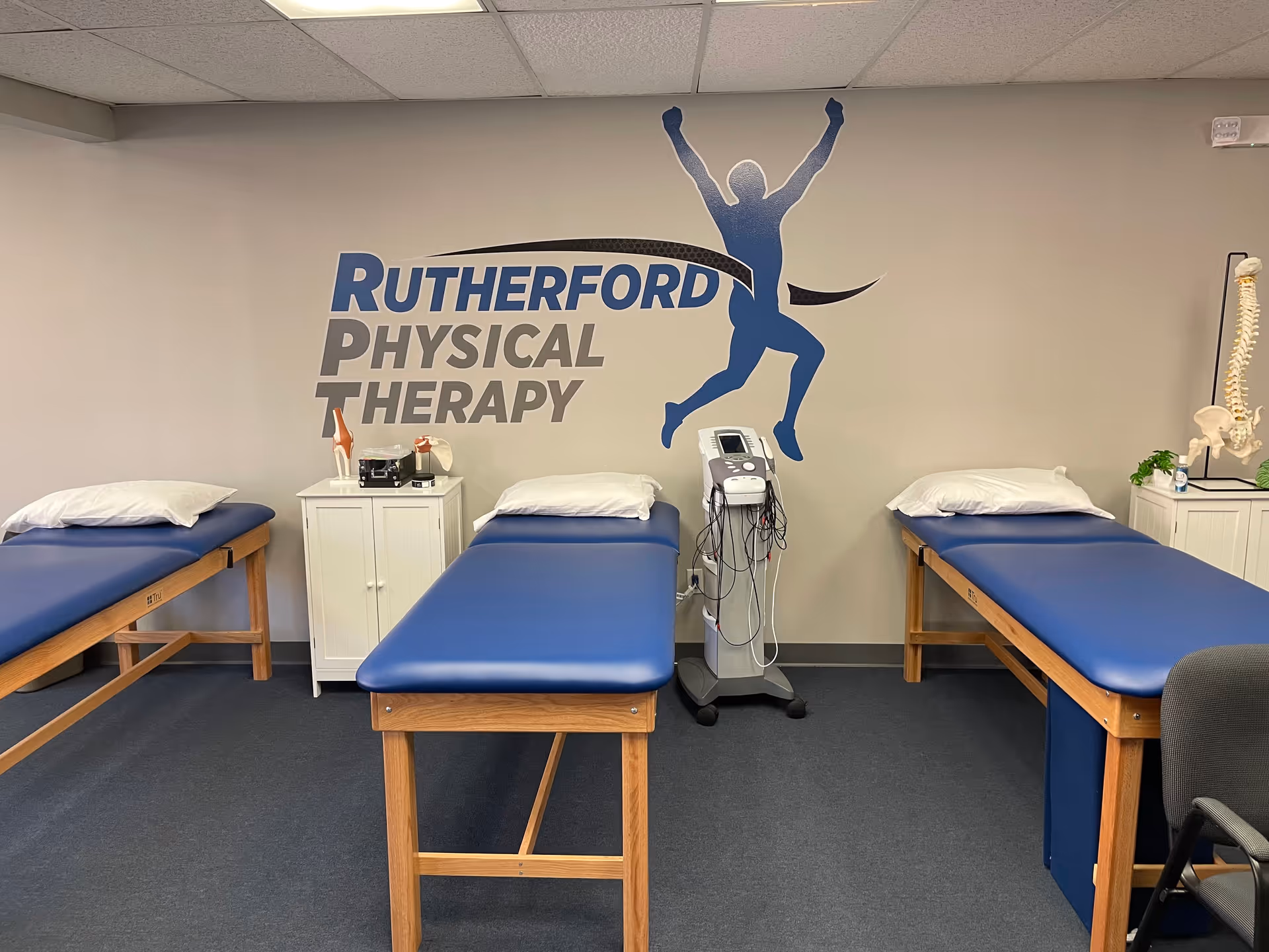 A Rutherford Physical Therapy treatment room with three blue treatment tables, equipment, and a wall mural.