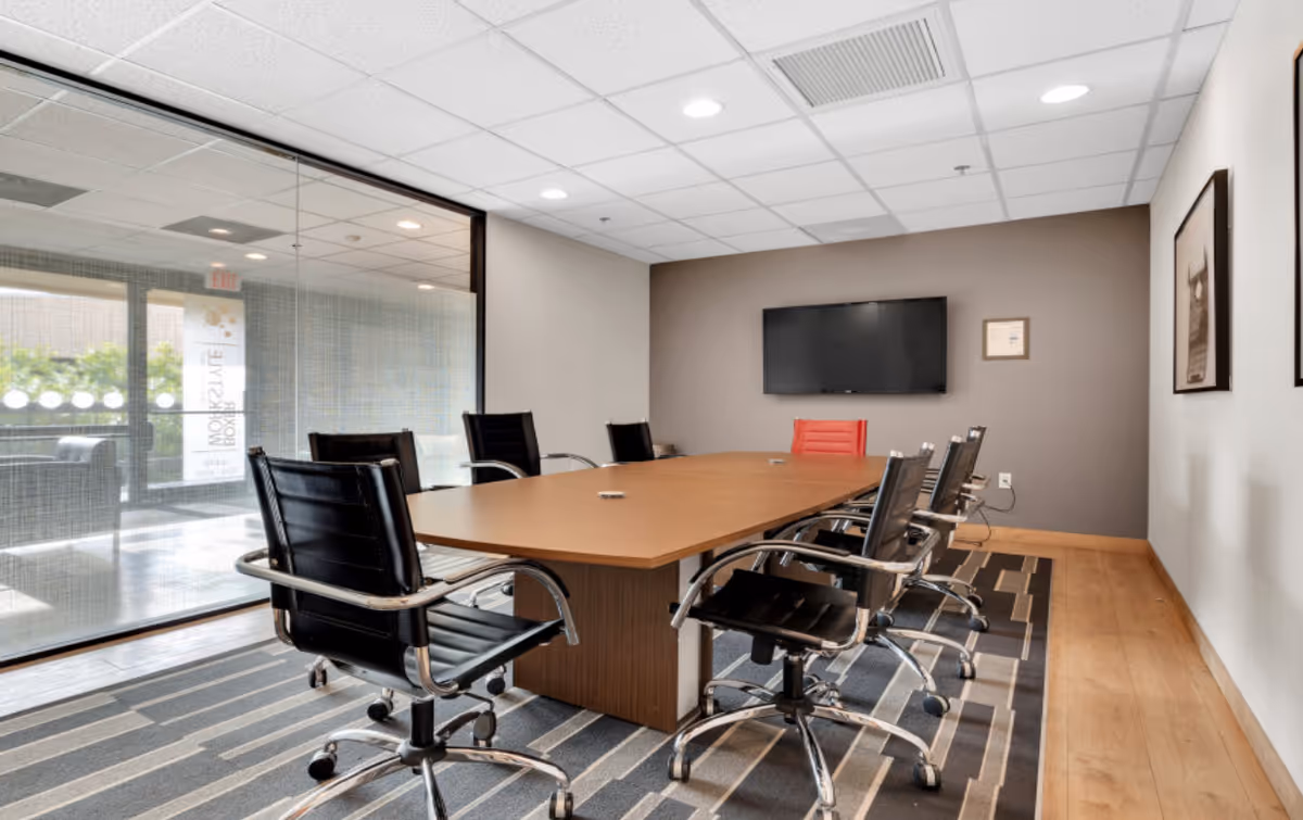 Modern conference room featuring a large wooden table, black leather chairs, glass partition, and TV.