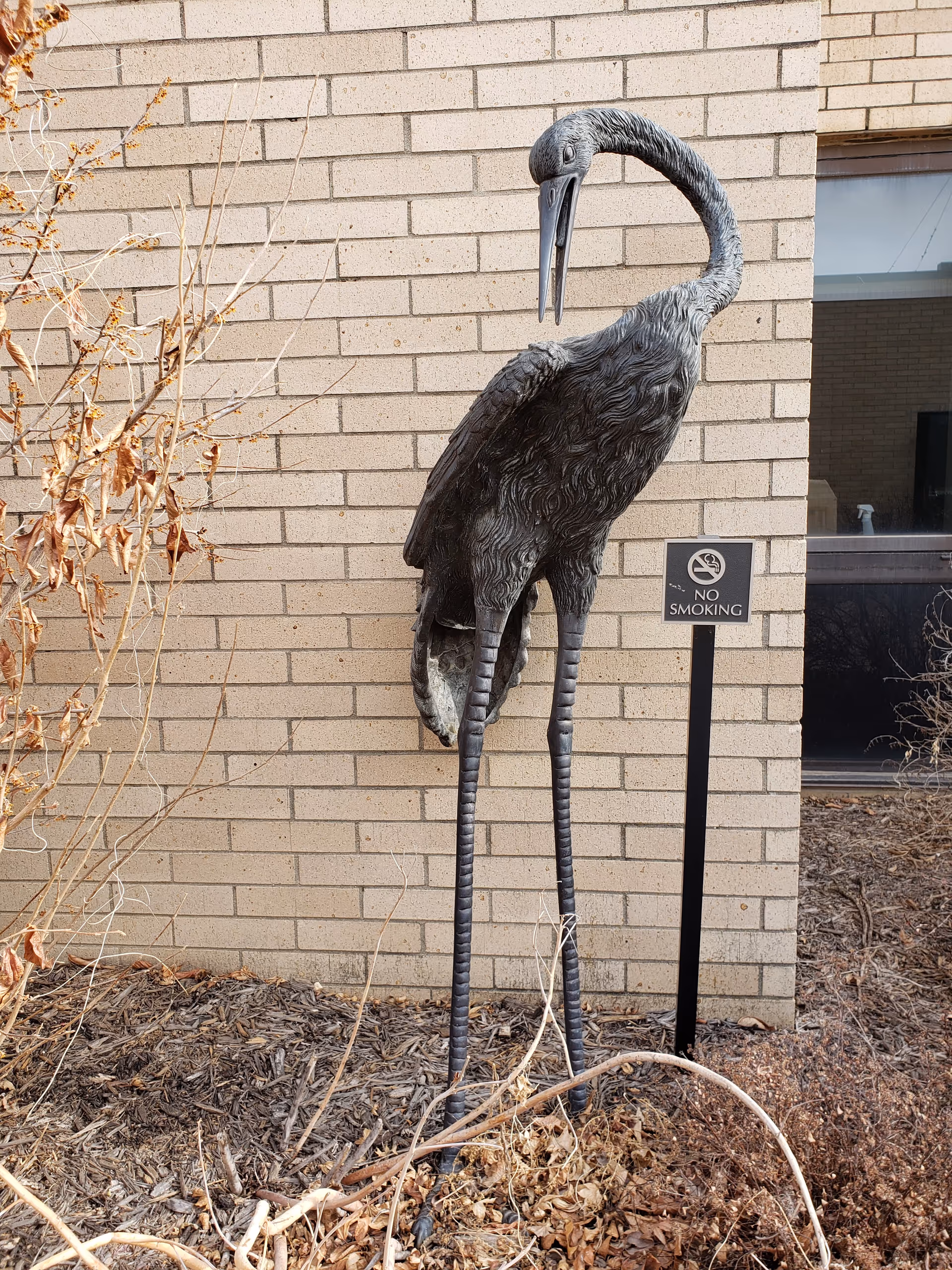 Metal bird sculpture stands outside a brick building beside a no-smoking sign.
