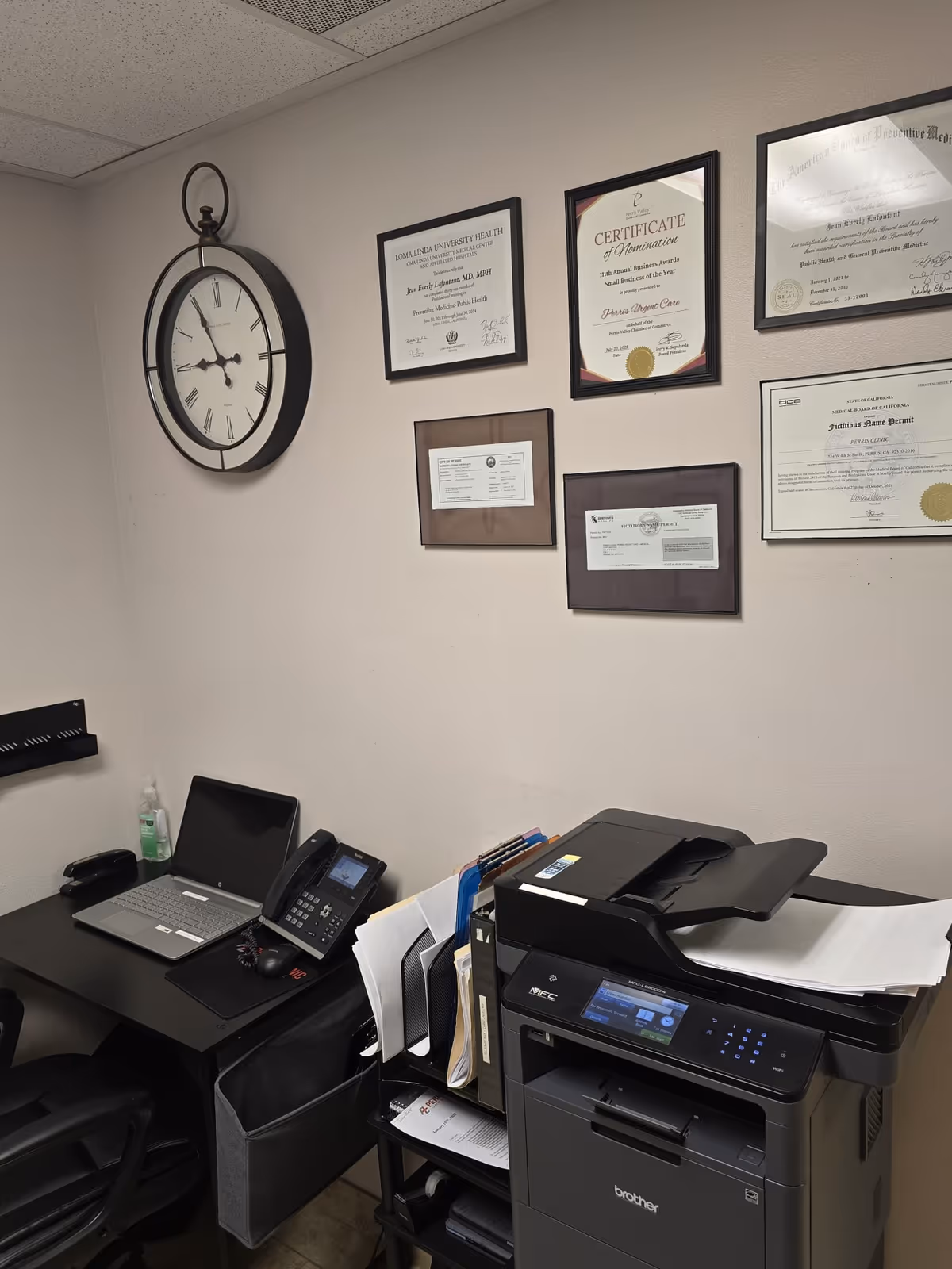 Office corner with wall clock, framed certificates, desk laptop, phone, and a multifunction printer.