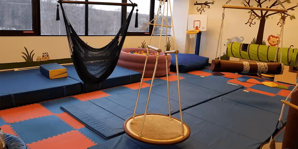 Indoor sensory gym with padded blue mats, a hanging hammock, ropes, and play equipment.