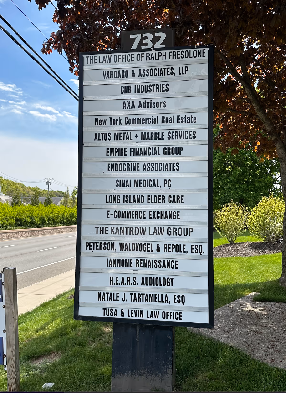 Tall outdoor directory sign listing multiple businesses along a street with trees nearby.