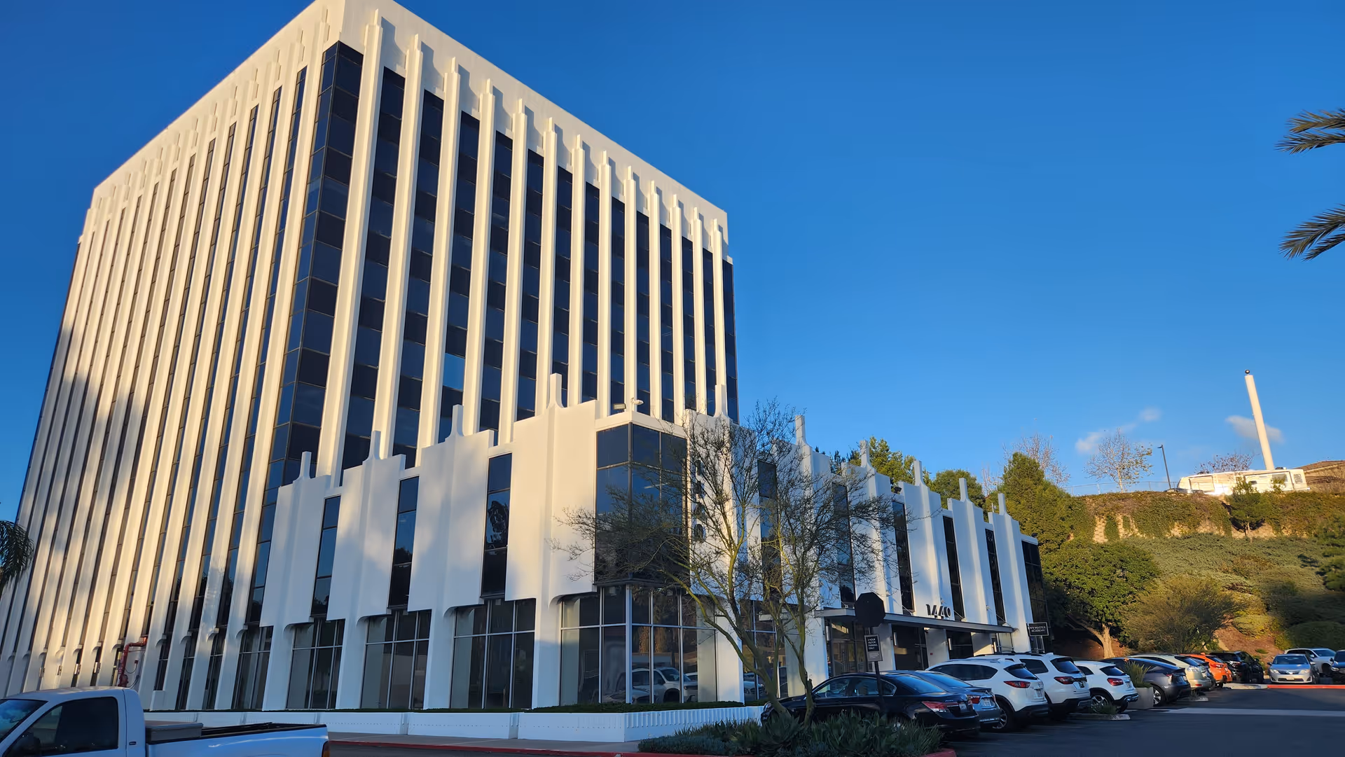 Modern white multi-story building with glass windows and rows of parked cars under blue sky.
