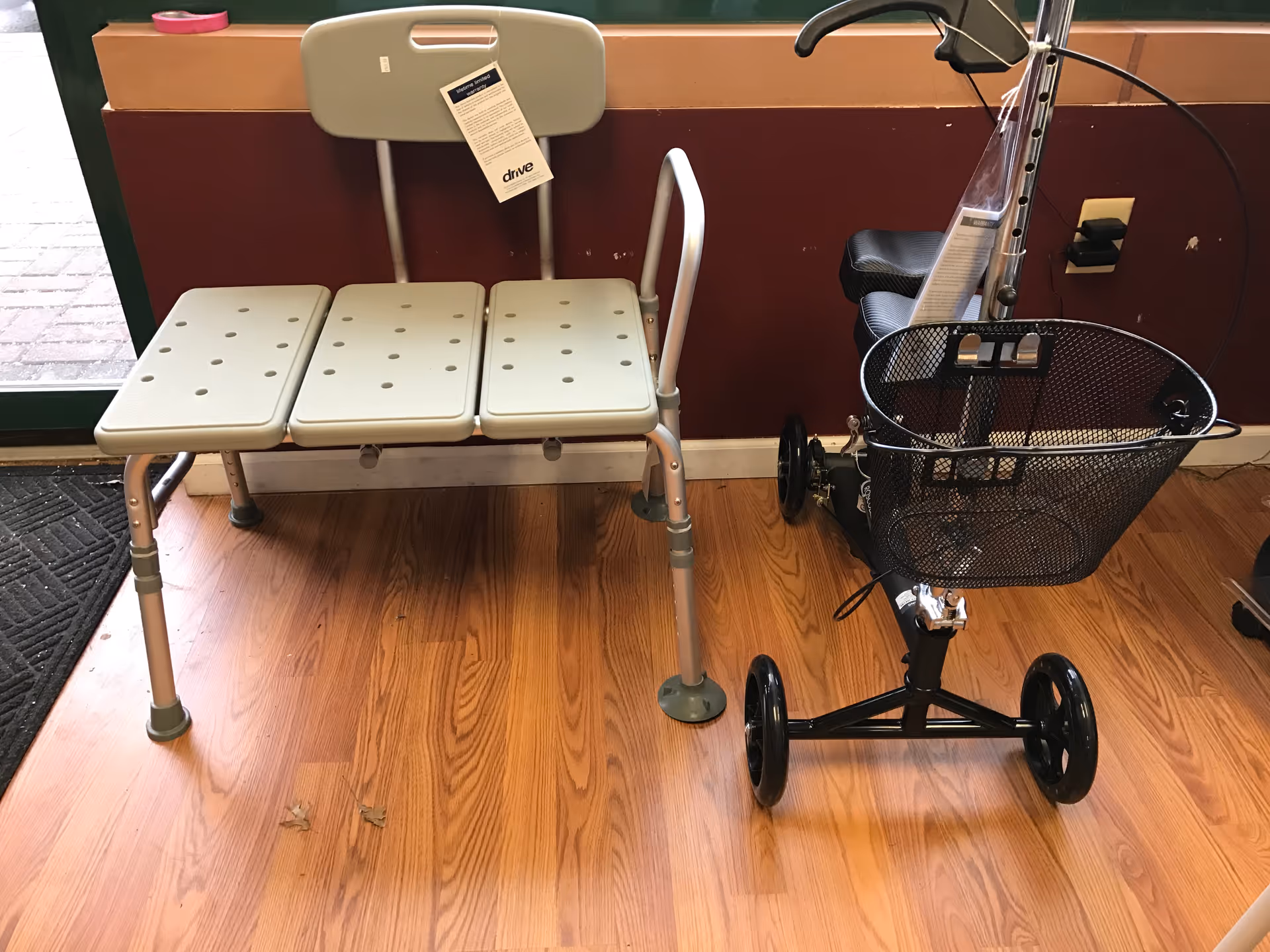 Mobility aids, including a walker with a basket and a three-seat bathing bench, against a wall.