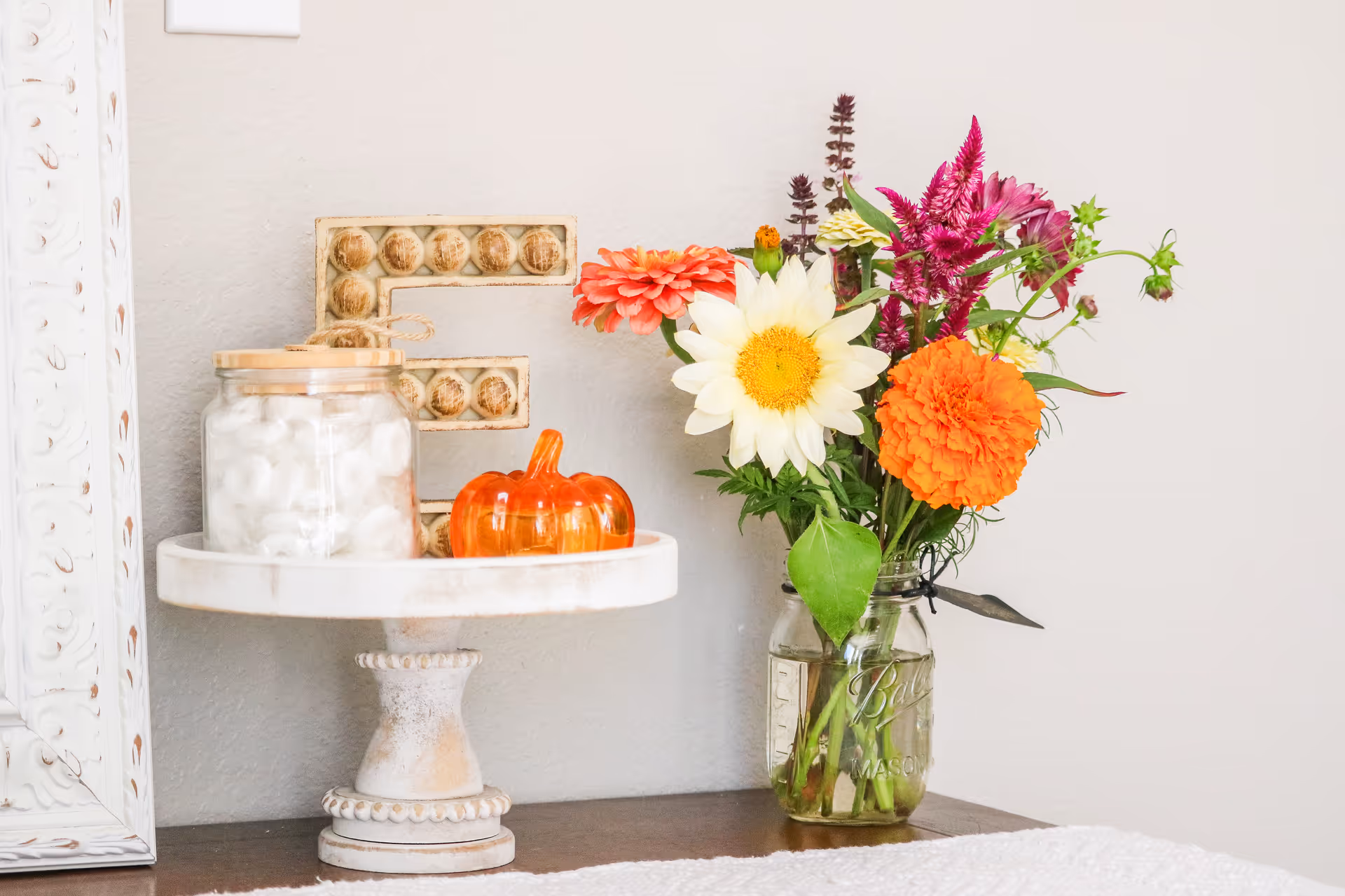 A white pedestal table displays a mason jar bouquet, pumpkin, and jars of treats.