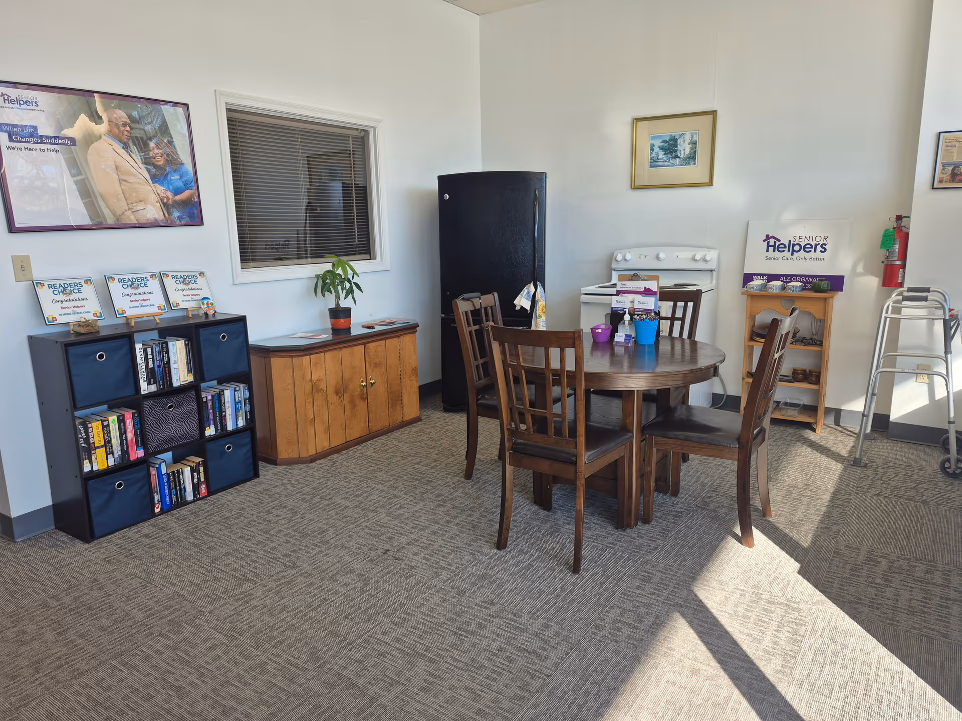 Common area with round table, chairs, bookshelf, cabinet, fridge, and a walker nearby.