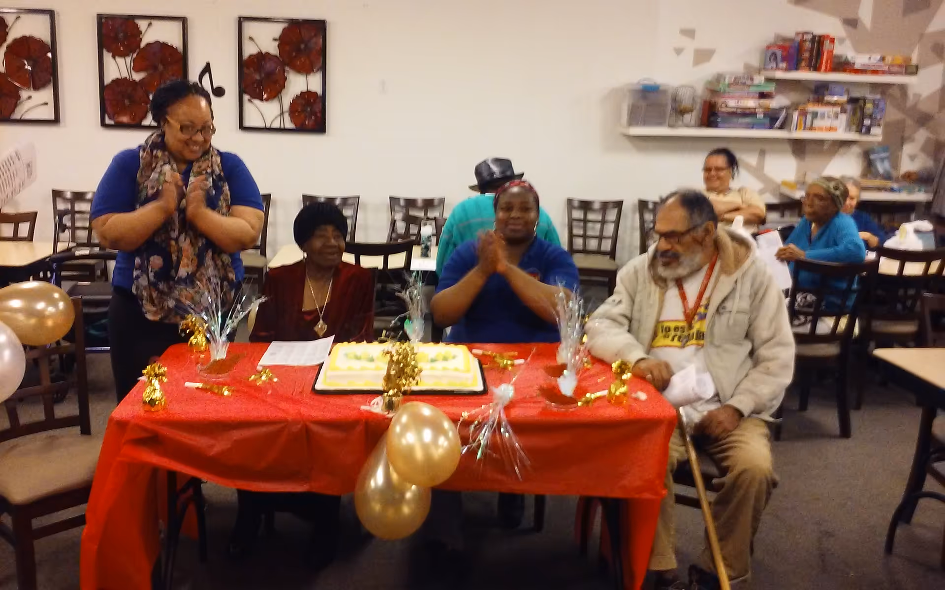 Group of seniors gathered around a decorated cake at a dining room table.
