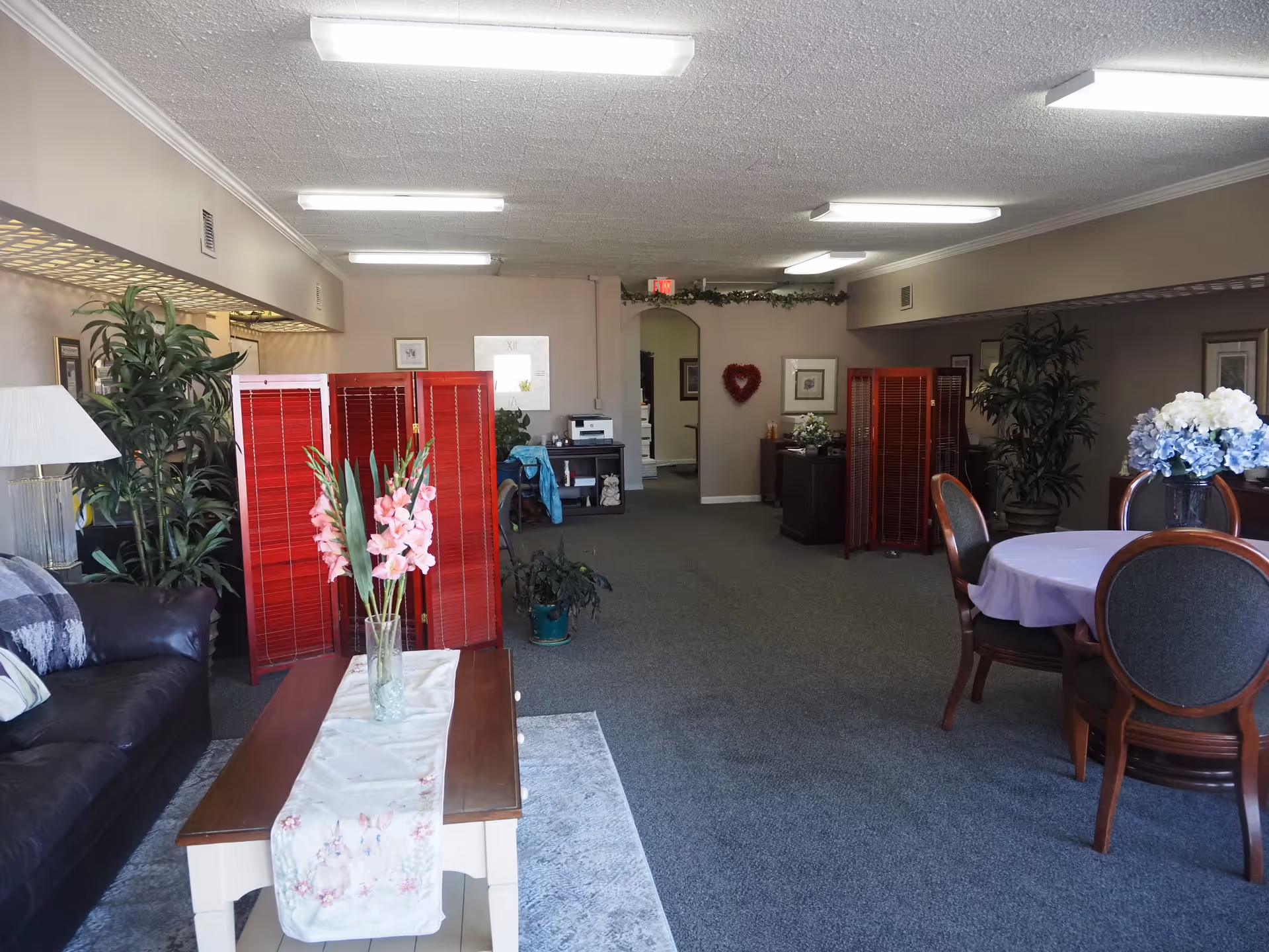 Senior living common area with sofa, coffee table, flowers, red room dividers, and a dining table.
