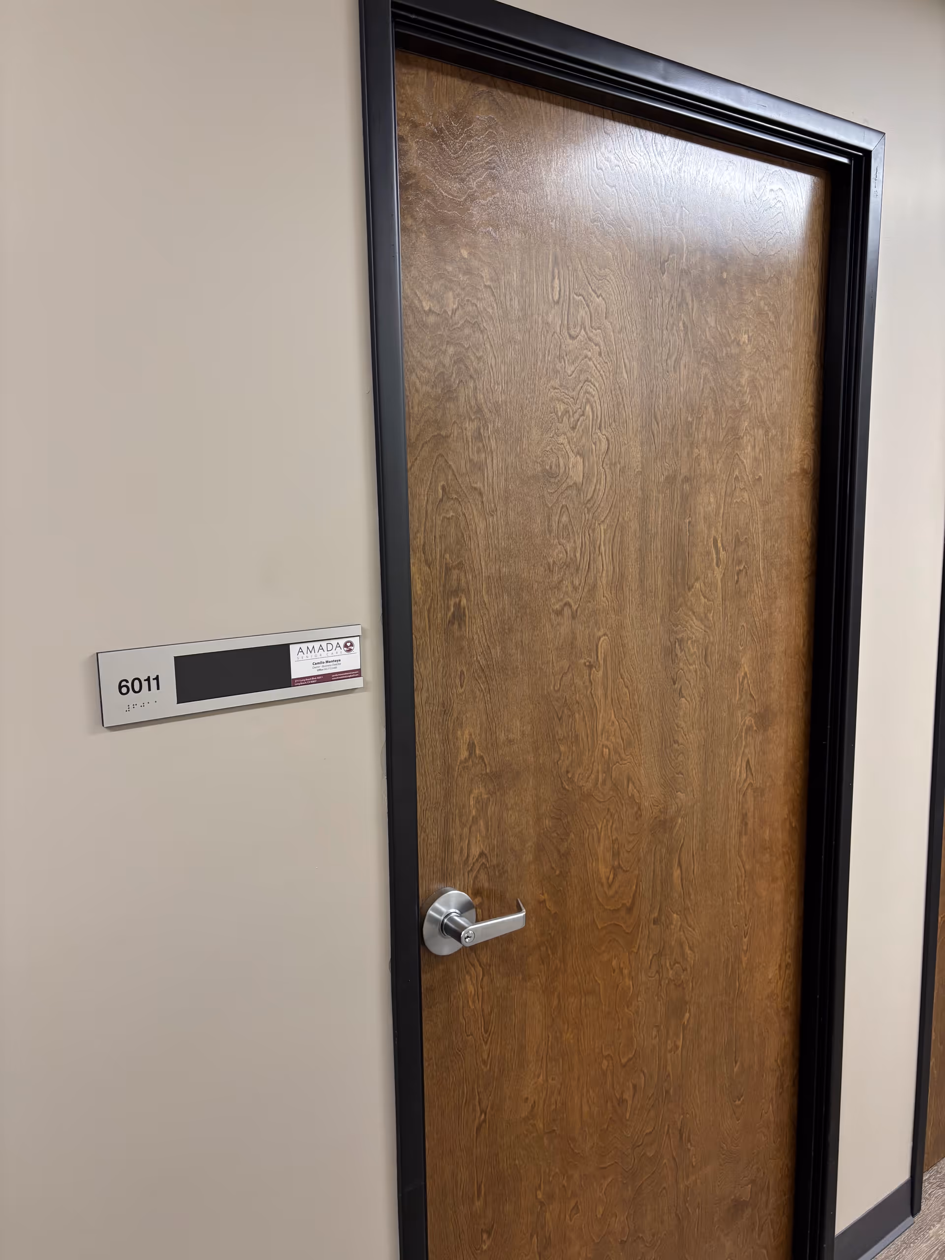 Wooden interior door with a metal handle in a beige hallway beside a room number plaque.