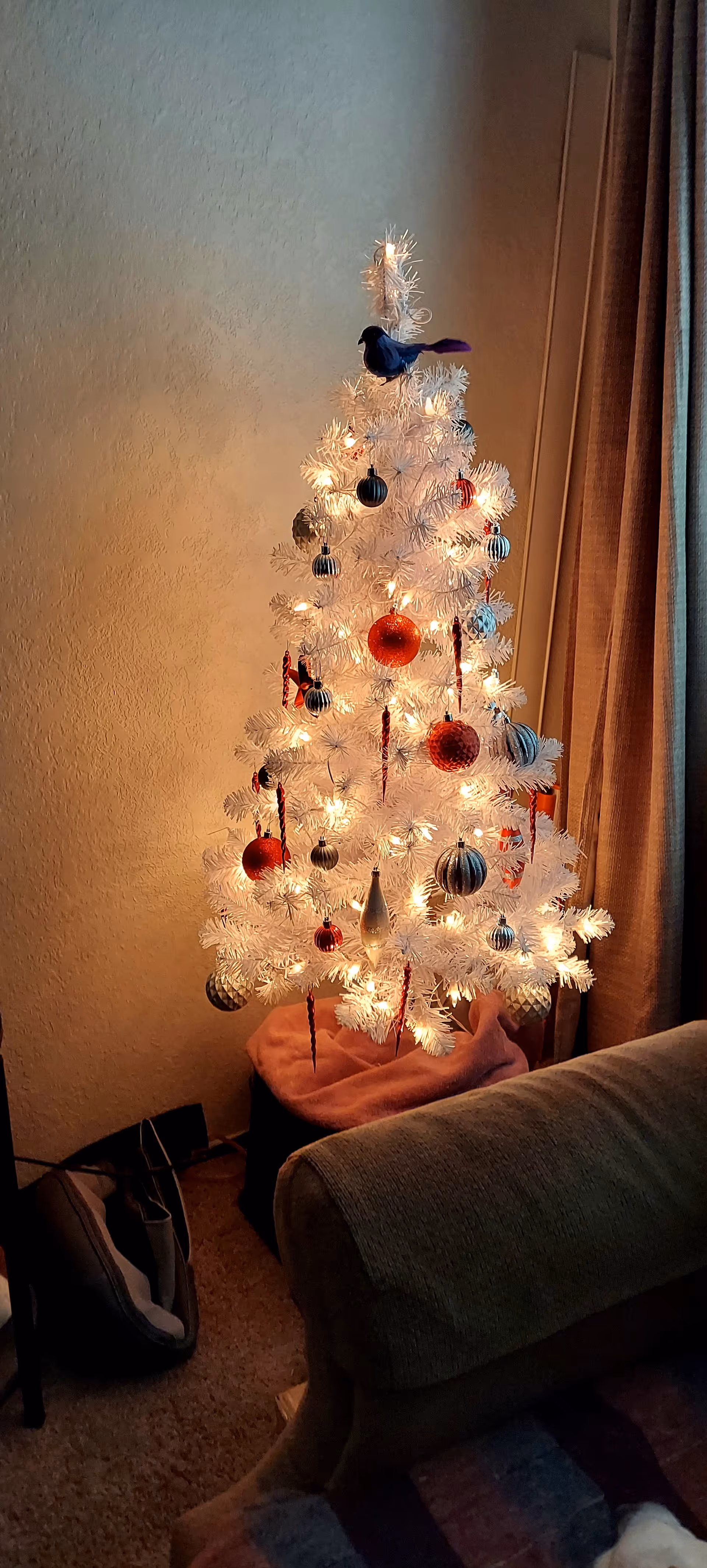 A white Christmas tree with lights and ornaments sits beside a couch in a living room.