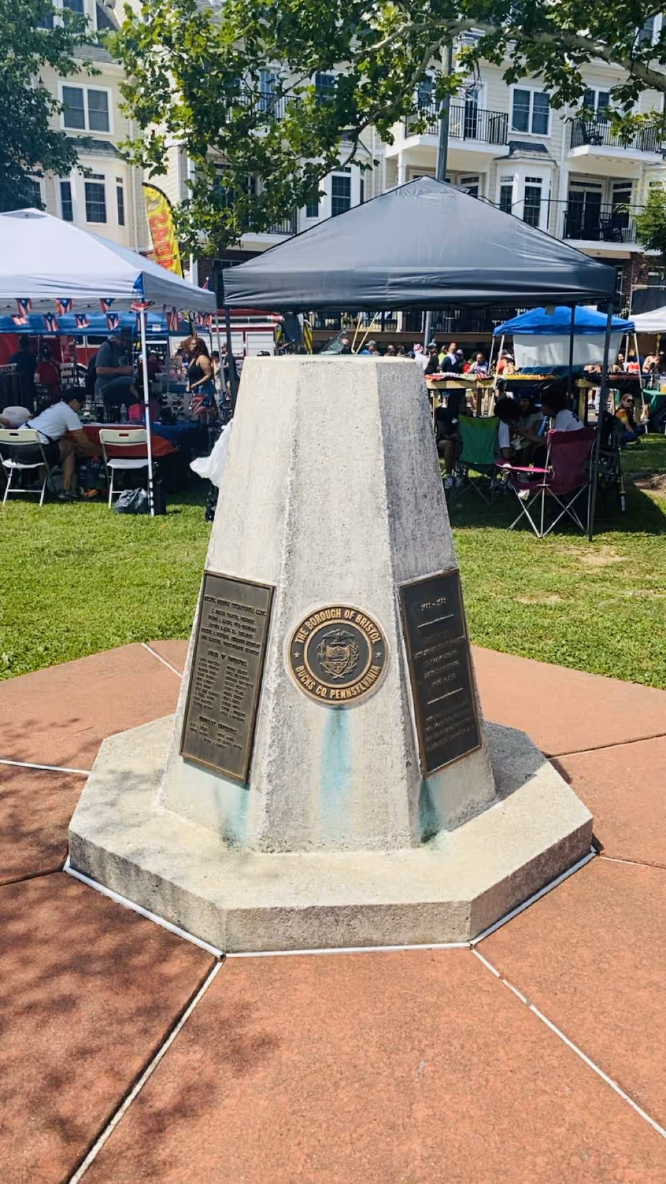 Outdoor park event with tents, attendees, and a central obelisk monument.