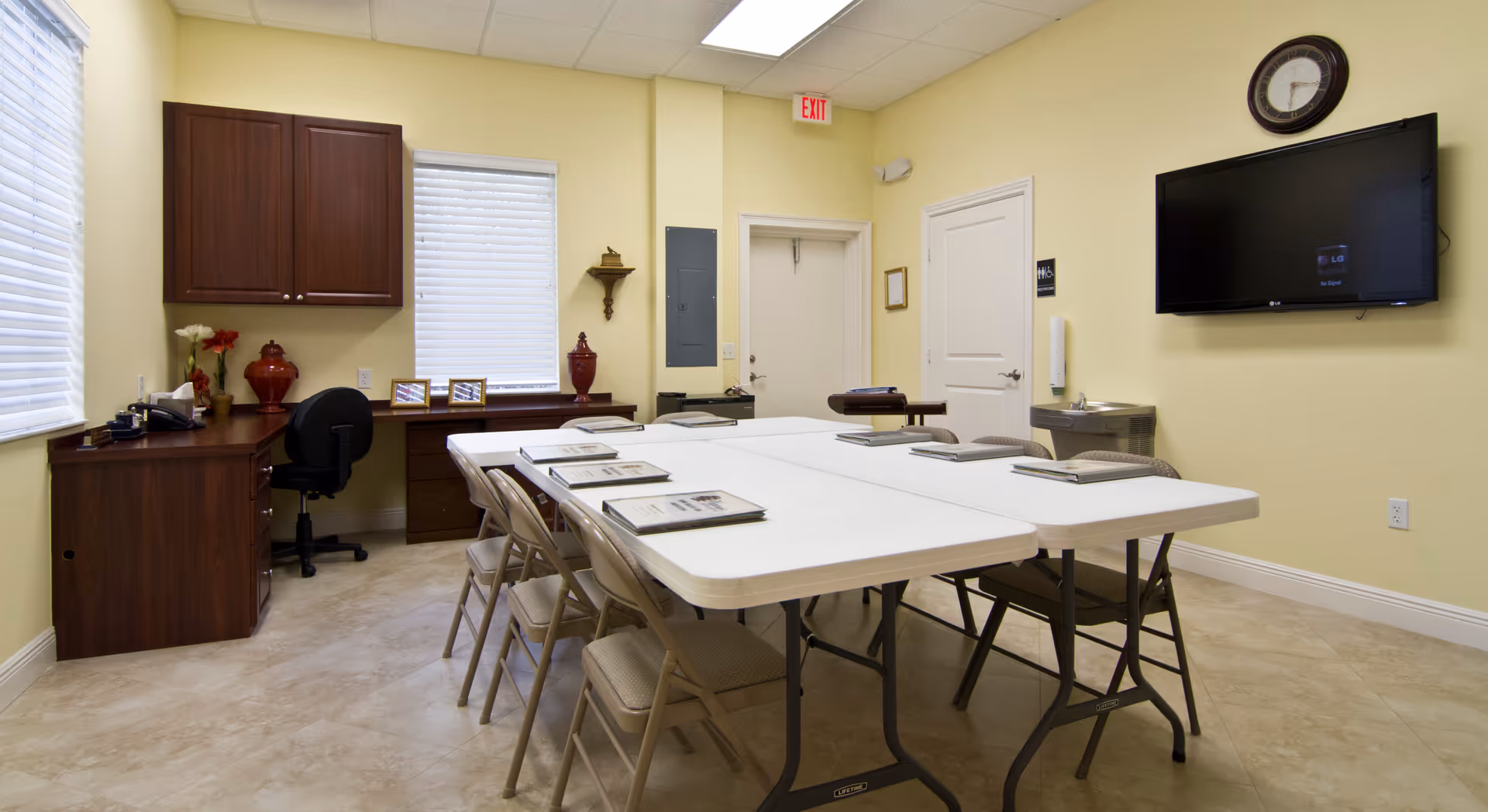 Bright institutional meeting room with folding tables, chairs, cabinetry, and a wall-mounted TV.