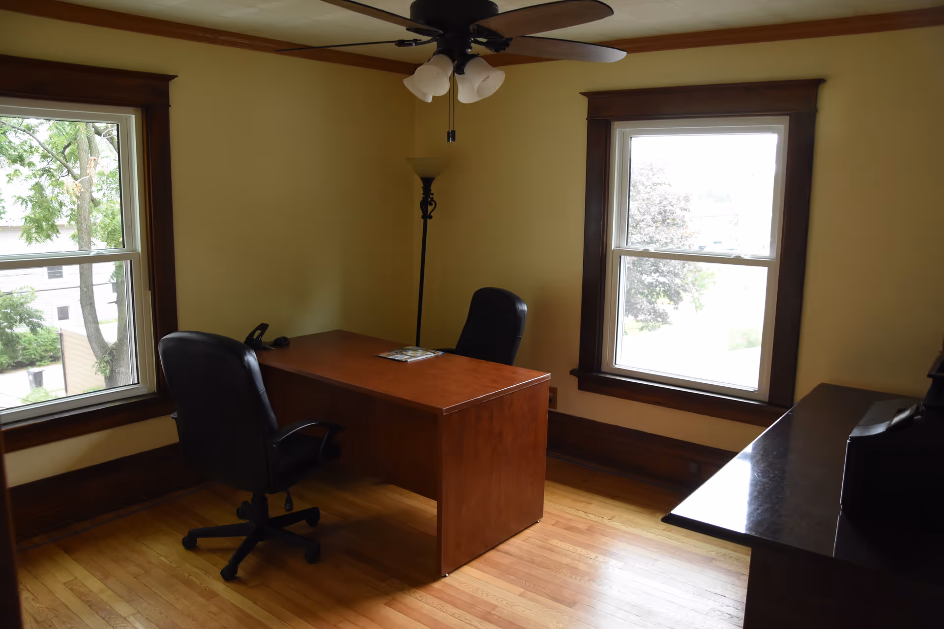 A small office with a wooden desk, two chairs, and two windows in a bright room.